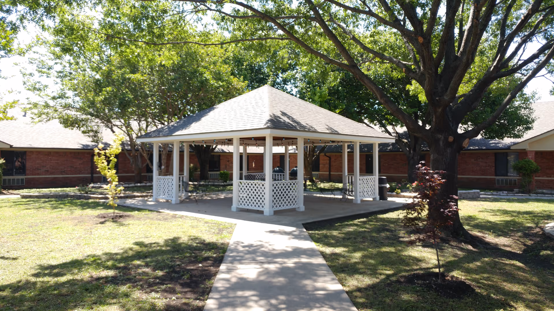 Sunlit white gazebo in a grassy courtyard with a brick building in the background.
