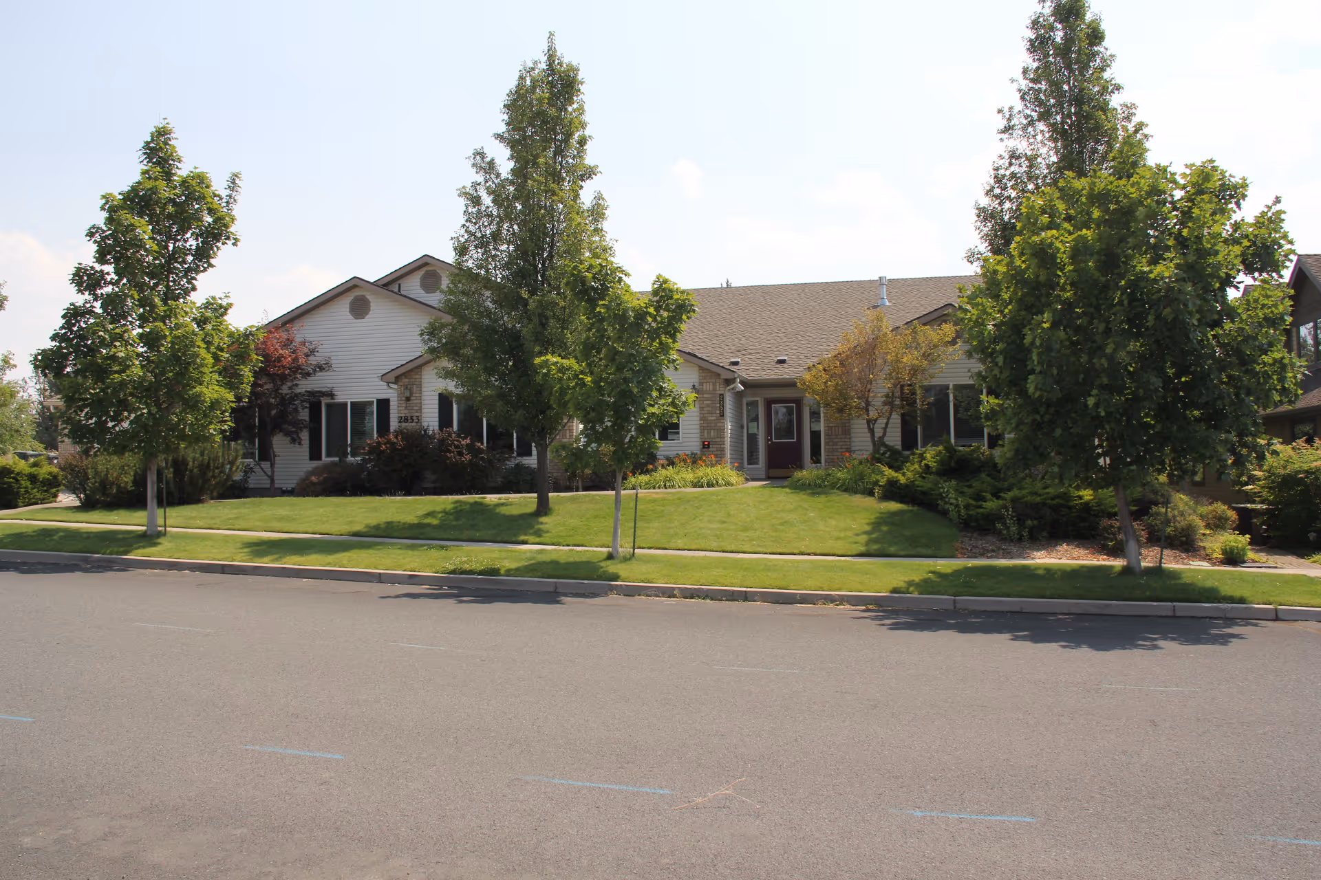 Front view of a single-story residential building with a lawn, trees, and a sidewalk along the street.