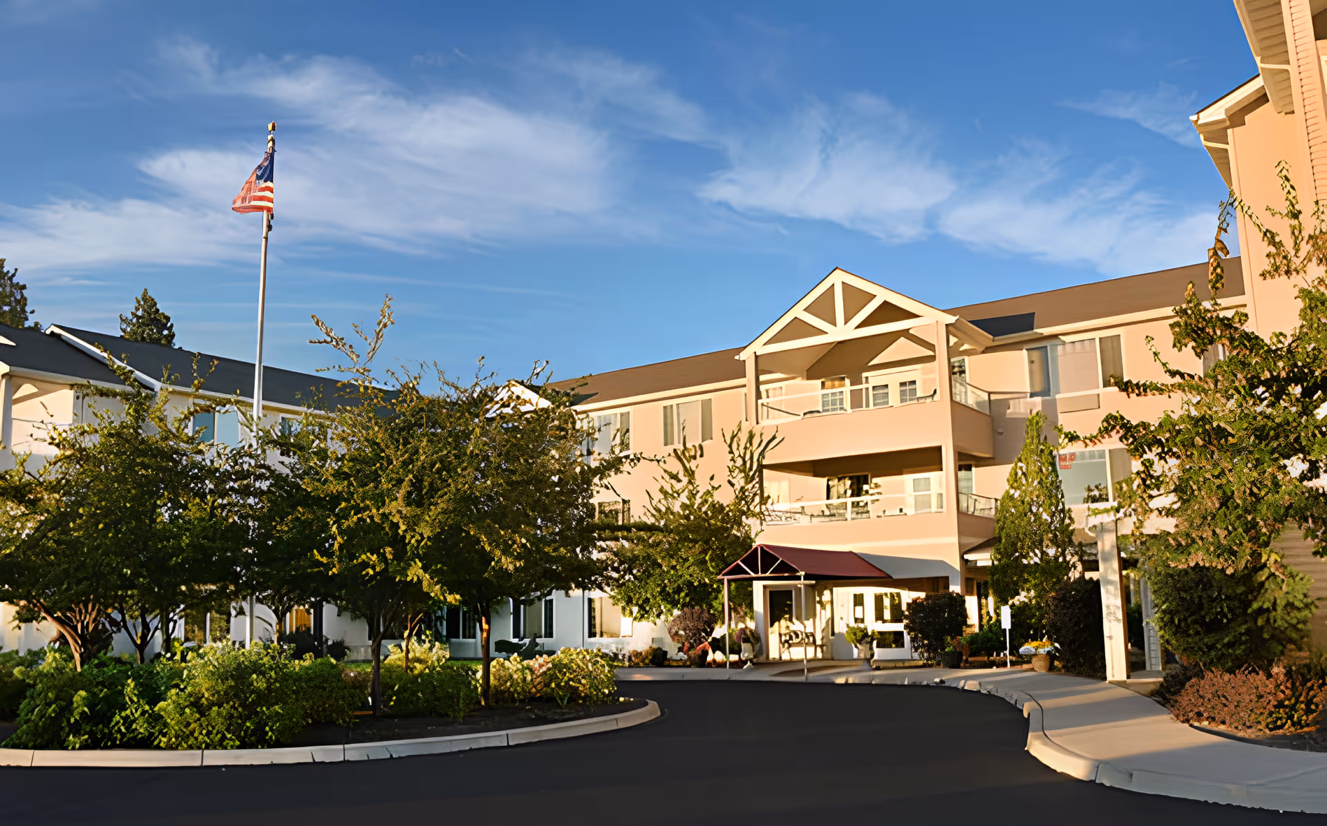 Front exterior of a multi-story senior living building with a covered entrance, landscaping, and an American flag.