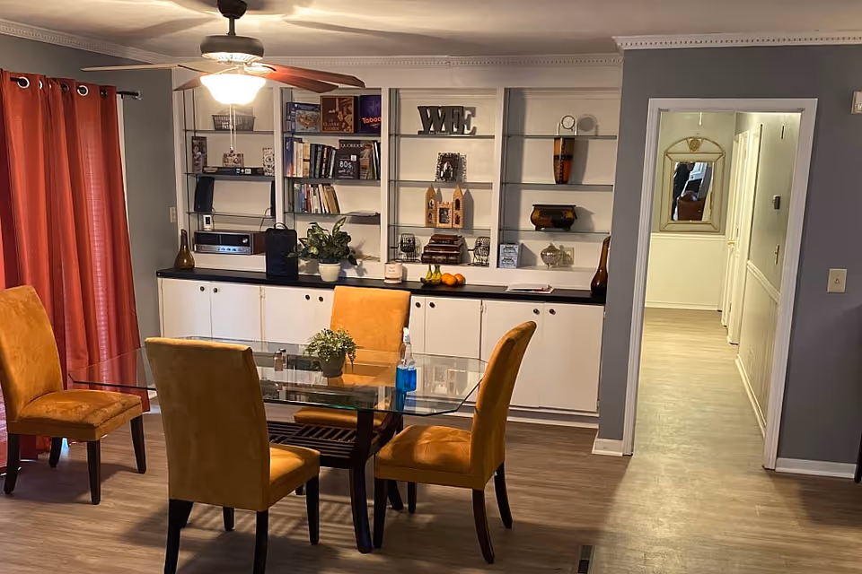 A dining area with a glass-top table surrounded by four mustard yellow upholstered chairs. Behind the table is a built-in white cabinet with shelves holding books, decorative items, and plants. Red curtains cover a window on the left side, and a ceiling fan with a light is above the table. A hallway with light-colored flooring and a mirror on the wall is visible through an open doorway on the right.