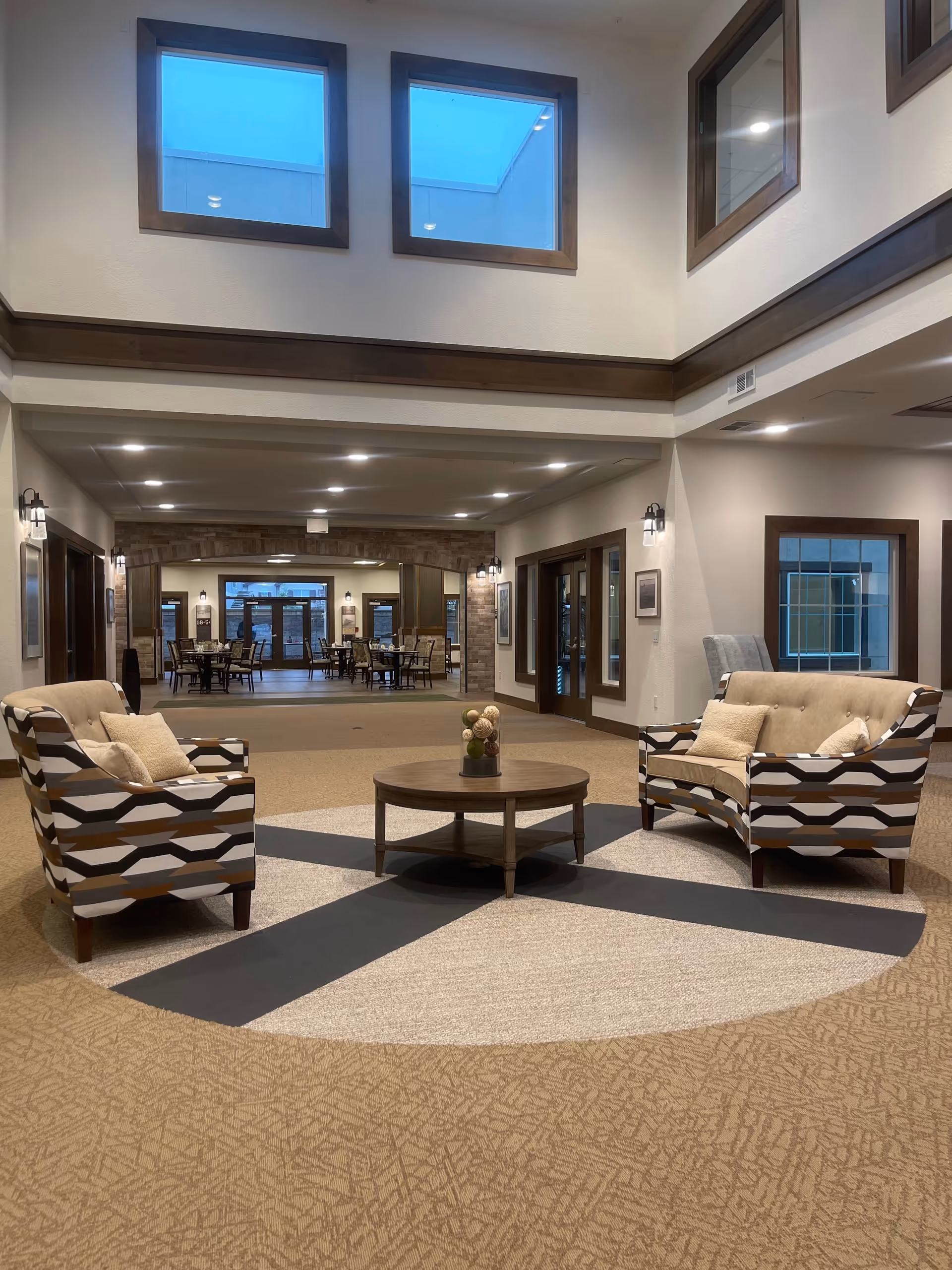 Lobby lounge with two patterned armchairs facing a round coffee table beneath skylights and a seating/dining area beyond.