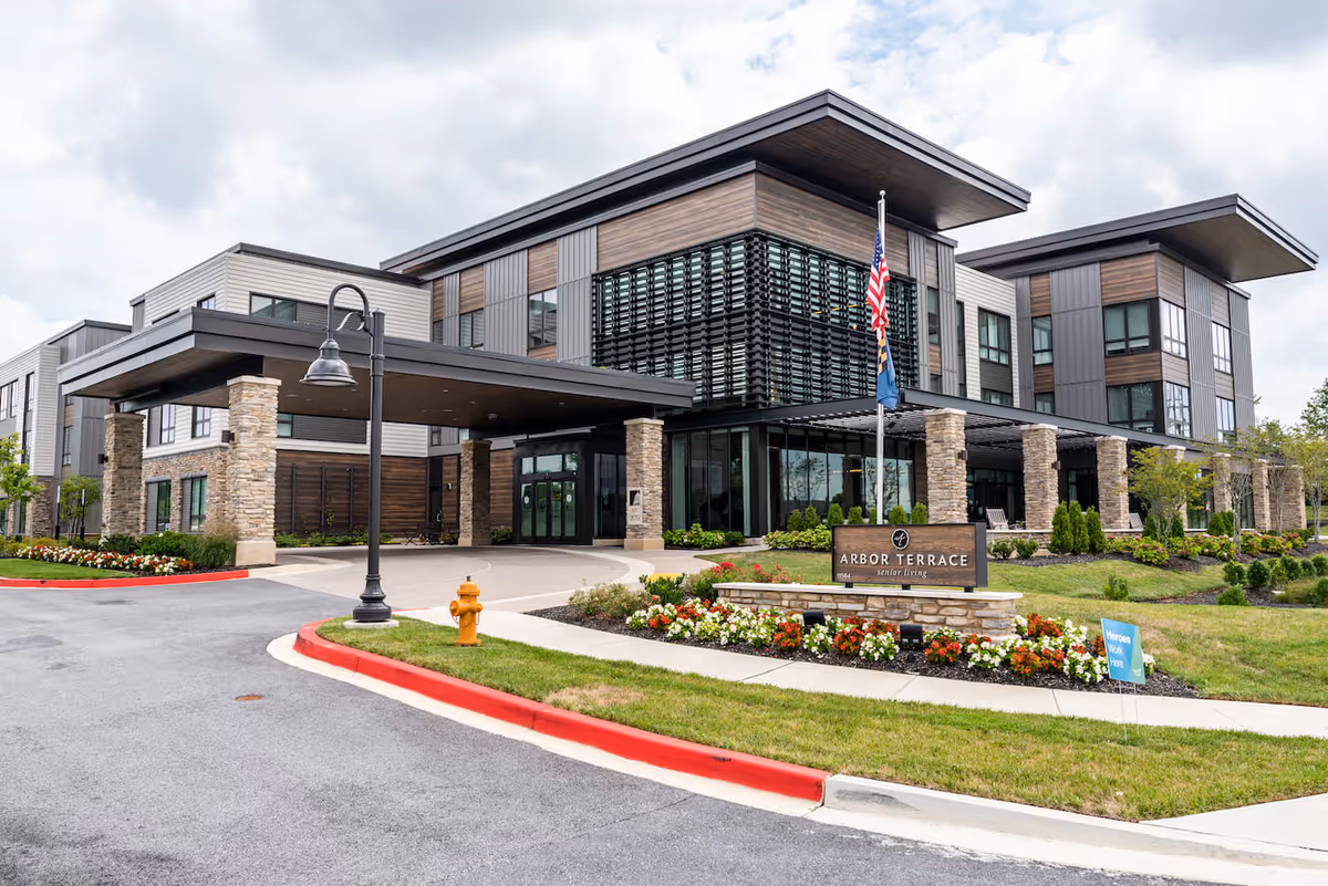 Exterior view of Arbor Terrace Fulton senior living facility showing a modern multi-story building with stone pillars, large windows, and a covered entrance. The landscaped area in front features colorful flowers, a flagpole with American and state flags, and a sign displaying the facility's name.