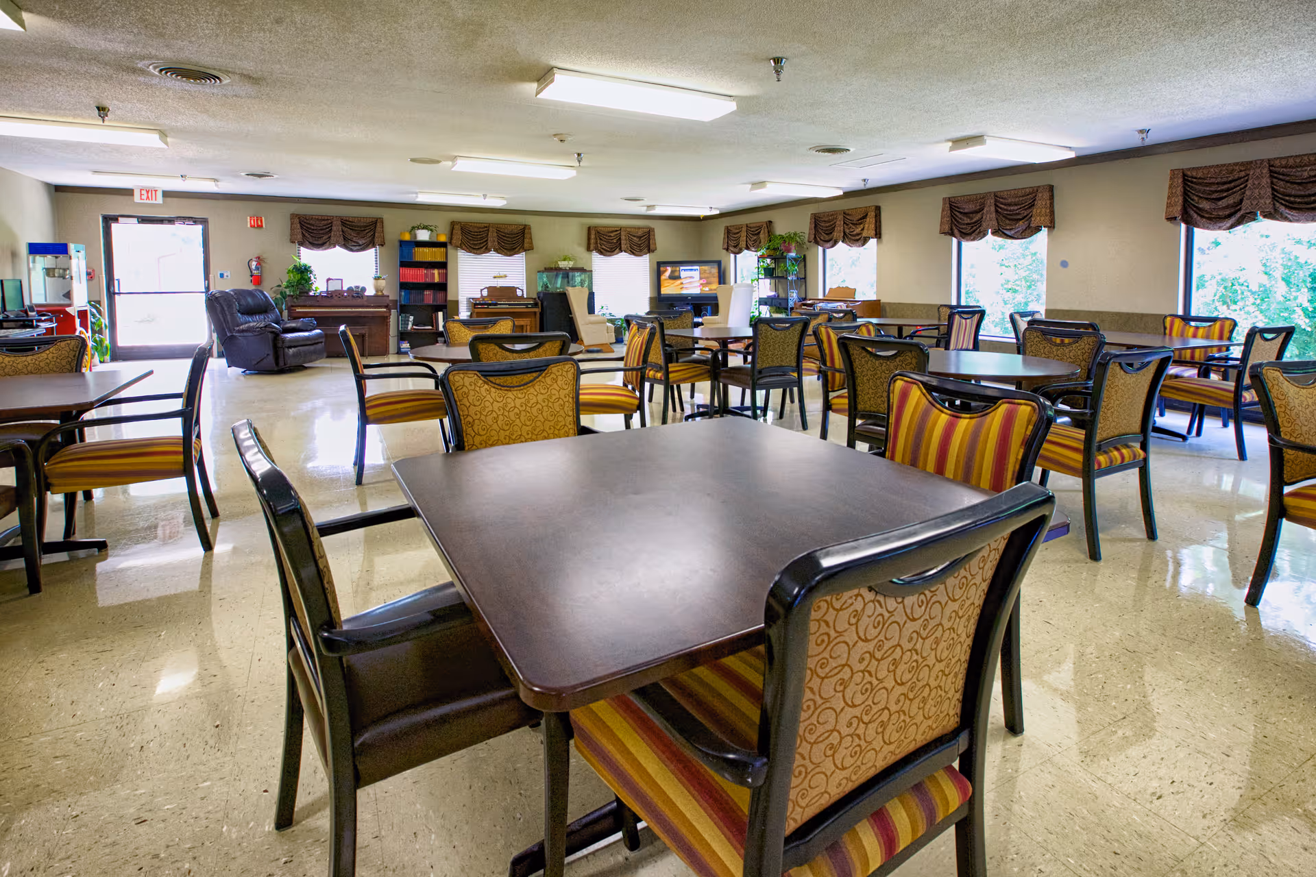 A spacious common area with multiple tables and chairs arranged for group seating. The chairs have patterned upholstery in shades of yellow and red. The room has several windows with brown valances letting in natural light. In the background, there is a television, a recliner chair, and some shelves with books and plants. The floor is a polished light-colored tile, and the ceiling has fluorescent lighting.