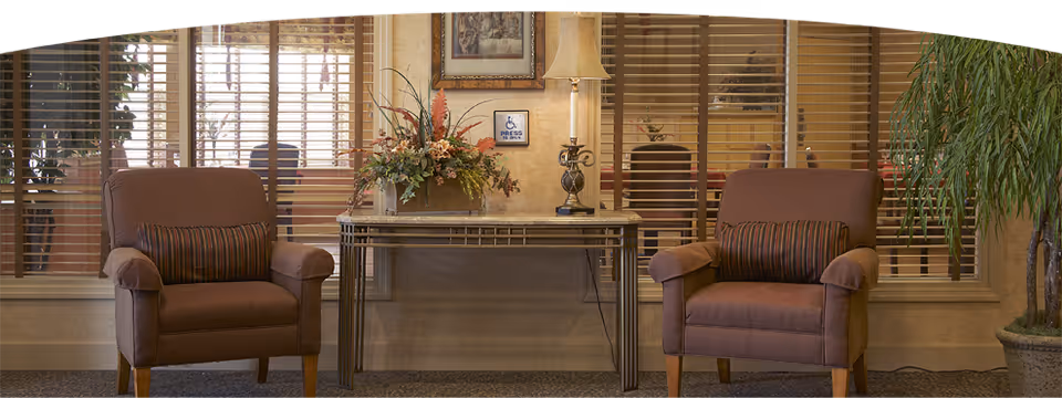 A cozy interior seating area with two brown upholstered armchairs, each with a striped cushion. Between the chairs is a narrow table with a floral arrangement and a decorative lamp. Behind the table is a wall with a framed picture and a small sign indicating accessibility. Large windows with wooden blinds are in the background, and a tall potted plant is visible on the right side.