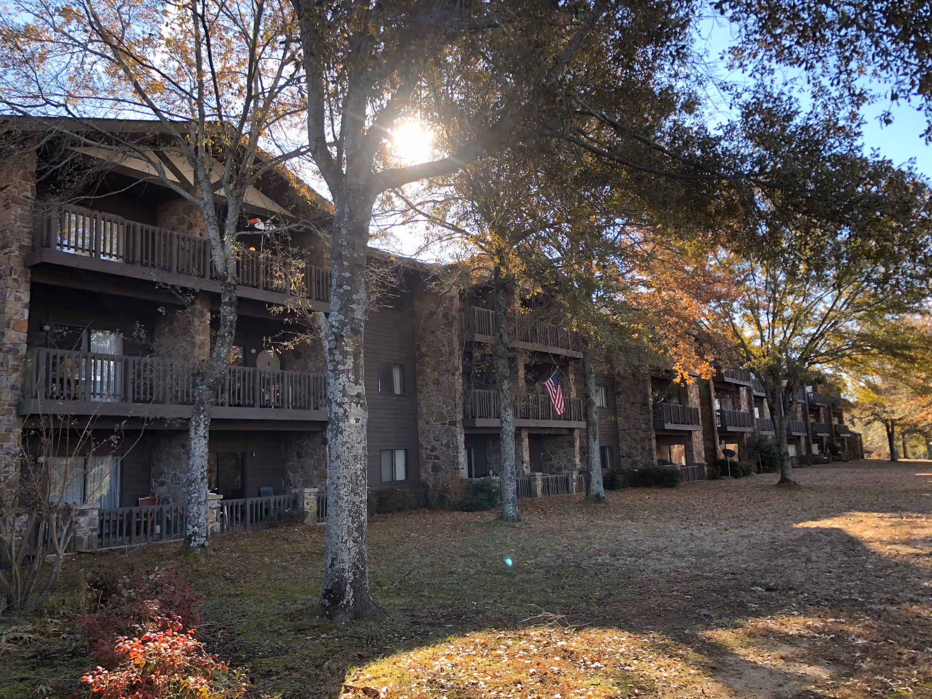Exterior view of a multi-story residential building with stone and wood facade, featuring balconies on each level. Trees with autumn foliage are in front of the building, and sunlight is shining through the branches.