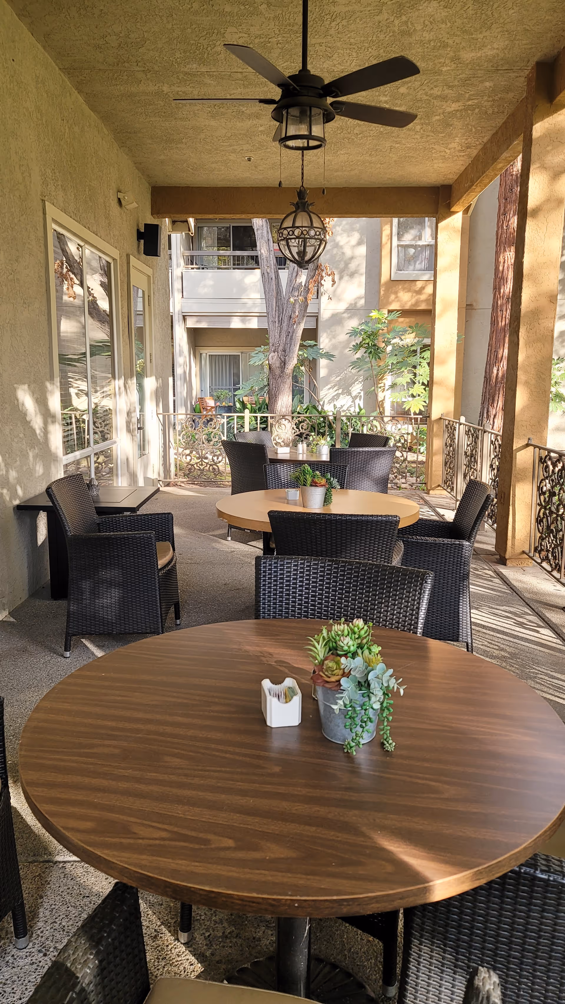Covered outdoor patio area with round wooden tables and black wicker chairs. Each table has a small potted succulent plant and a napkin holder. Ceiling fan and hanging light fixture are visible above. The patio overlooks a garden area with trees and plants, and there are balconies of an adjacent building in the background.
