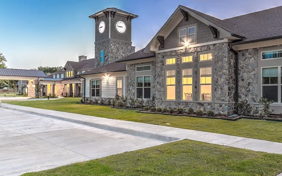 Exterior view of Ridgemere Senior Living facility at dusk, featuring a stone and siding building with large windows illuminated from inside, a clock tower, a covered entrance, and a well-maintained lawn and driveway.