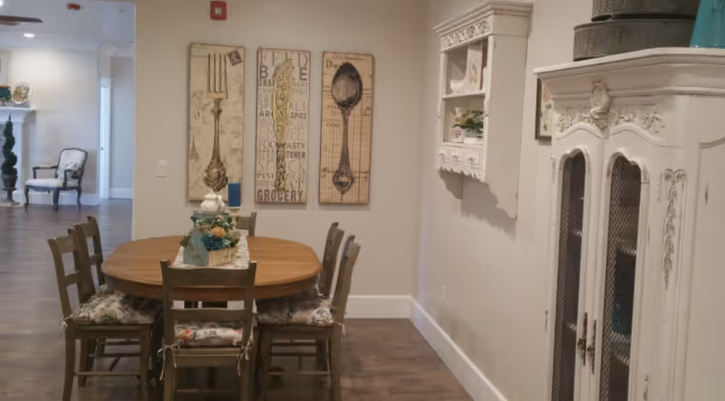 Round wooden dining table with six chairs and a centerpiece in a cozy common area with wall art and a white vintage cabinet.