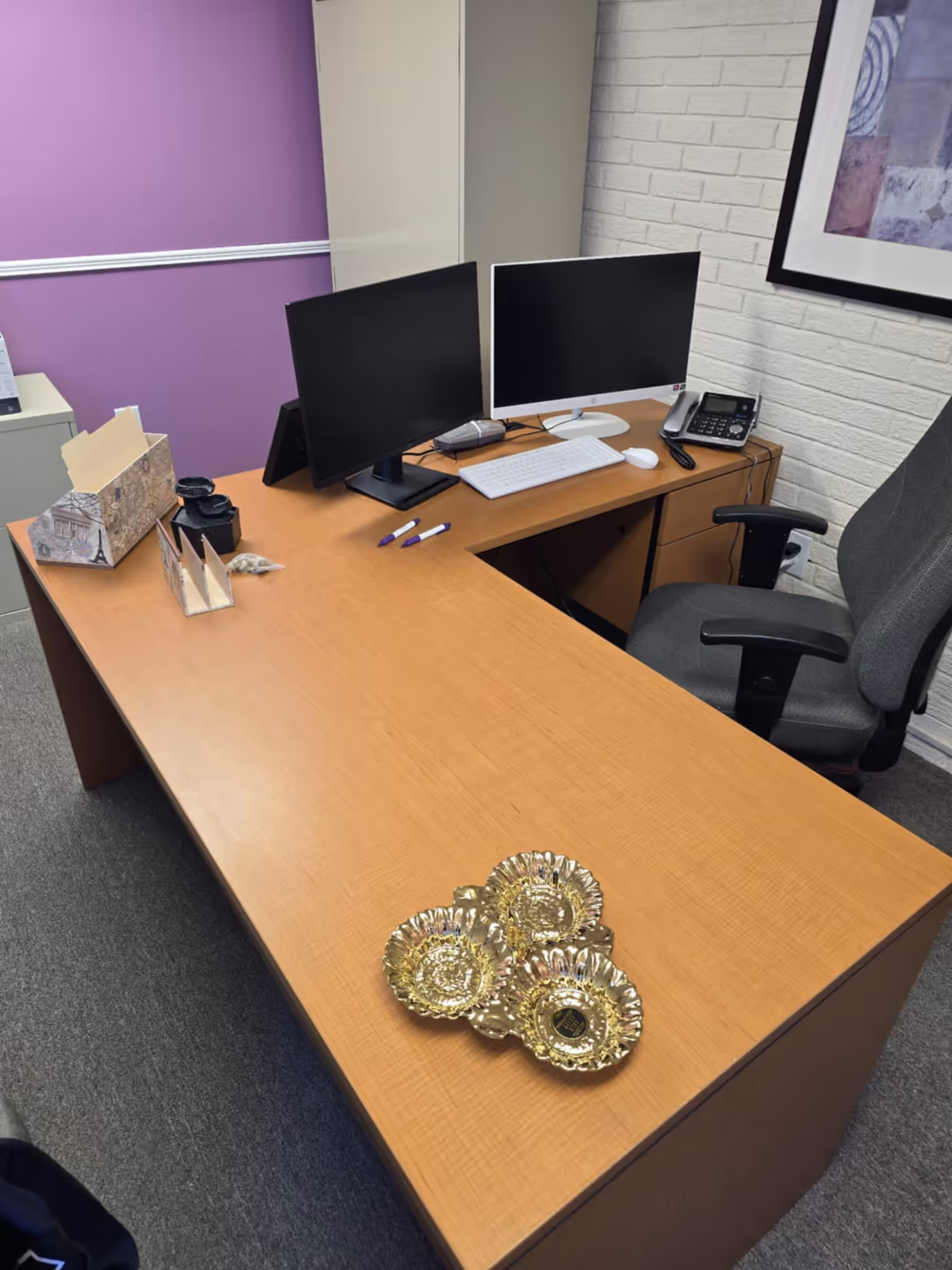 Office desk with dual monitors, keyboard, phone, and golden decorative coasters in a purple-walled room.