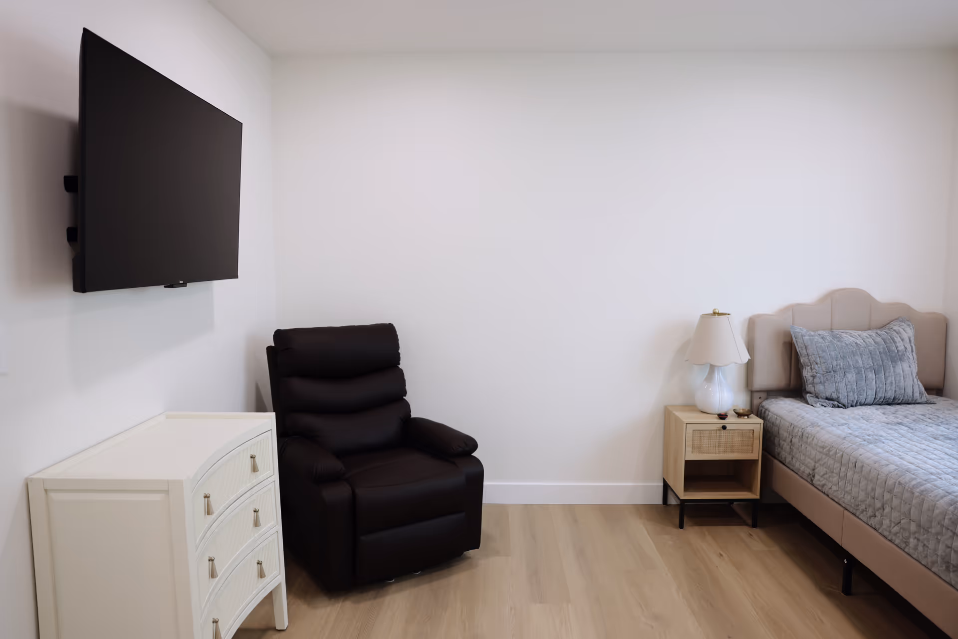 Minimalist bedroom with wall-mounted TV, beige bed, nightstand, lamp, and black recliner.