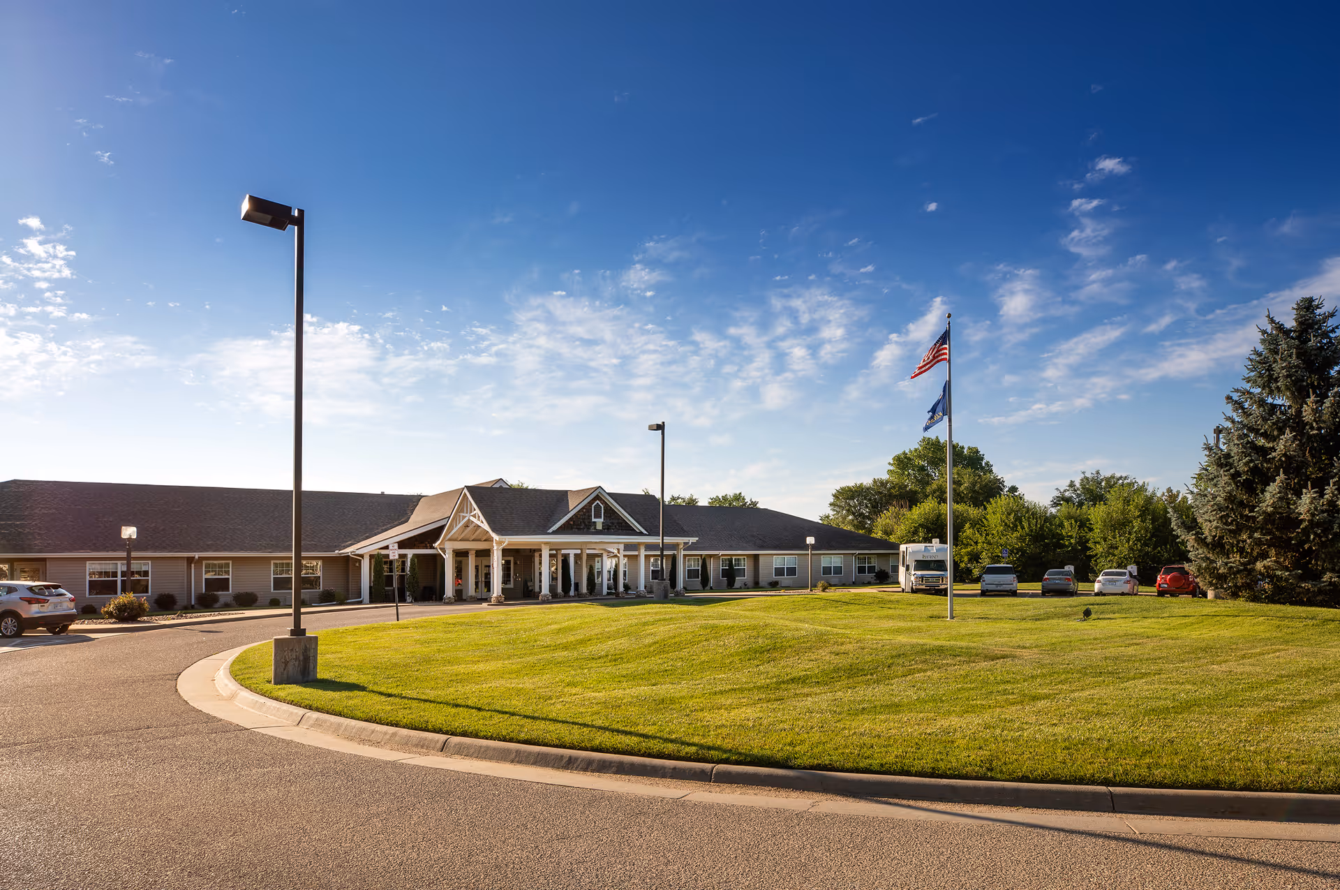 Exterior view of a single-story senior living facility building with a covered entrance, a curved driveway, a well-maintained lawn, a flagpole with the American flag and another flag, several parked cars, and a clear blue sky with some clouds.