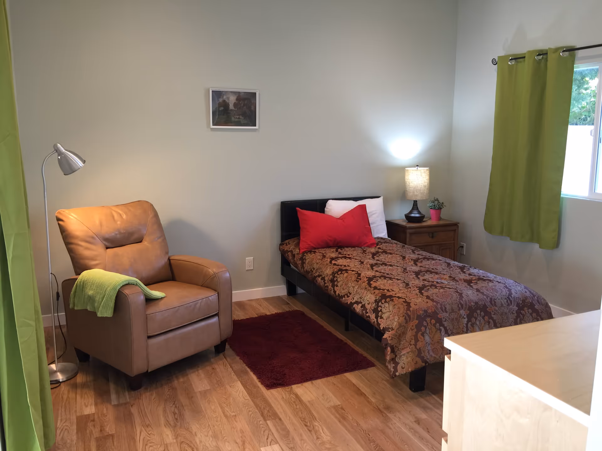 A cozy bedroom with a single bed covered in a patterned brown and orange bedspread, two red pillows, and a white pillow. Next to the bed is a wooden nightstand with a lamp and a small potted plant. A brown leather armchair with a green throw blanket is positioned near a floor lamp. The room has light-colored walls, wooden flooring, green curtains on the window, and a small red rug on the floor.