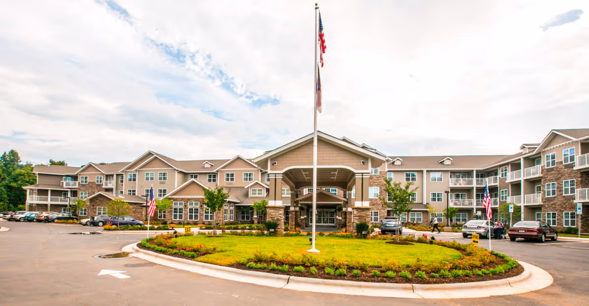 Front exterior view of a large, multi-story senior living facility with a covered entrance, landscaped roundabout with a flagpole, and several parked cars in the parking lot under a partly cloudy sky.