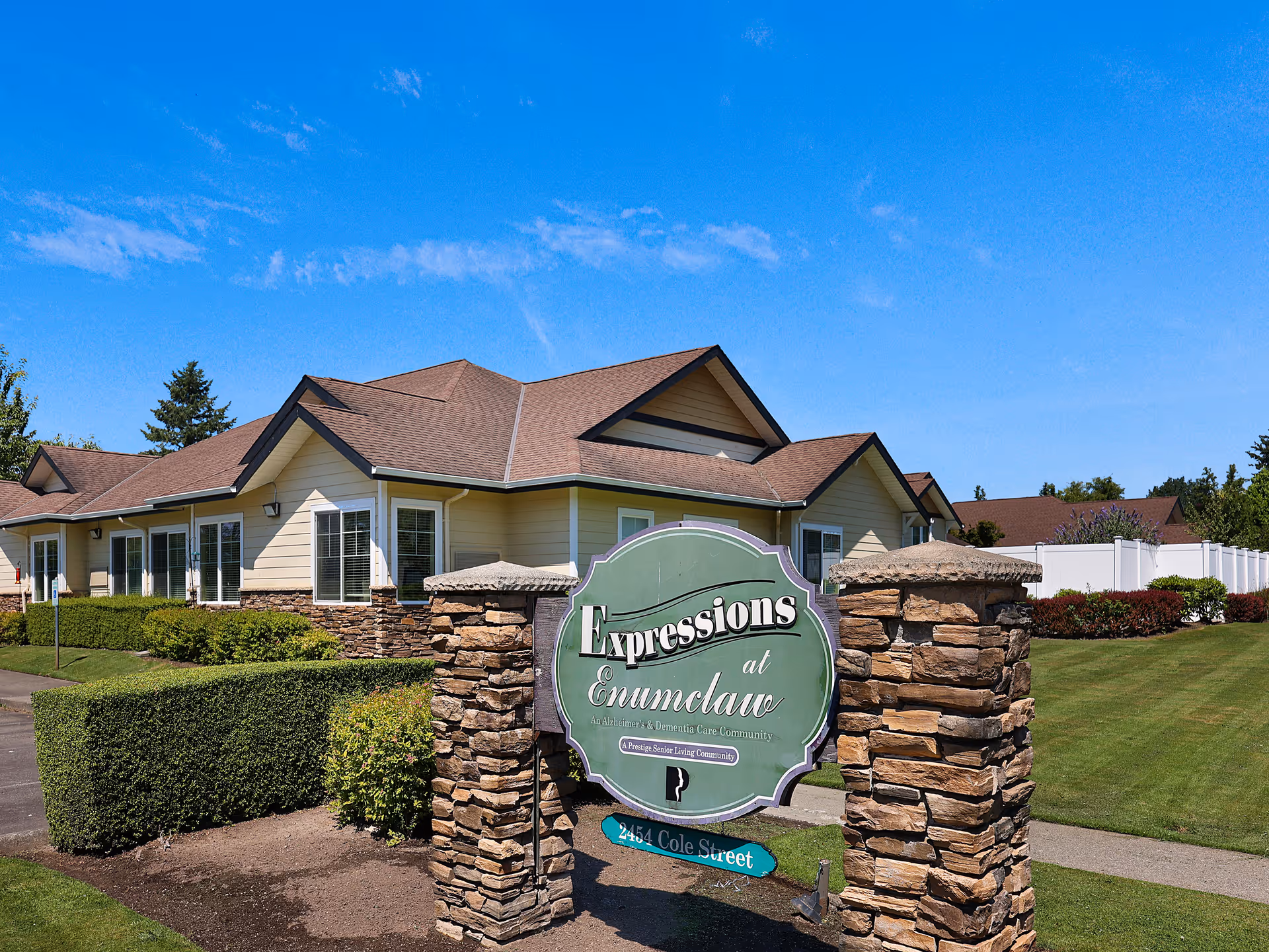 Exterior view of a single-story senior living facility building with beige siding and a brown roof under a clear blue sky. In front of the building is a stone pillar sign that reads 'Expressions at Enumclaw, An Alzheimer's & Dementia Care Community, A Prestige Senior Living Community, 2454 Cole Street.' The surrounding area includes well-maintained green lawns, bushes, and a paved walkway.