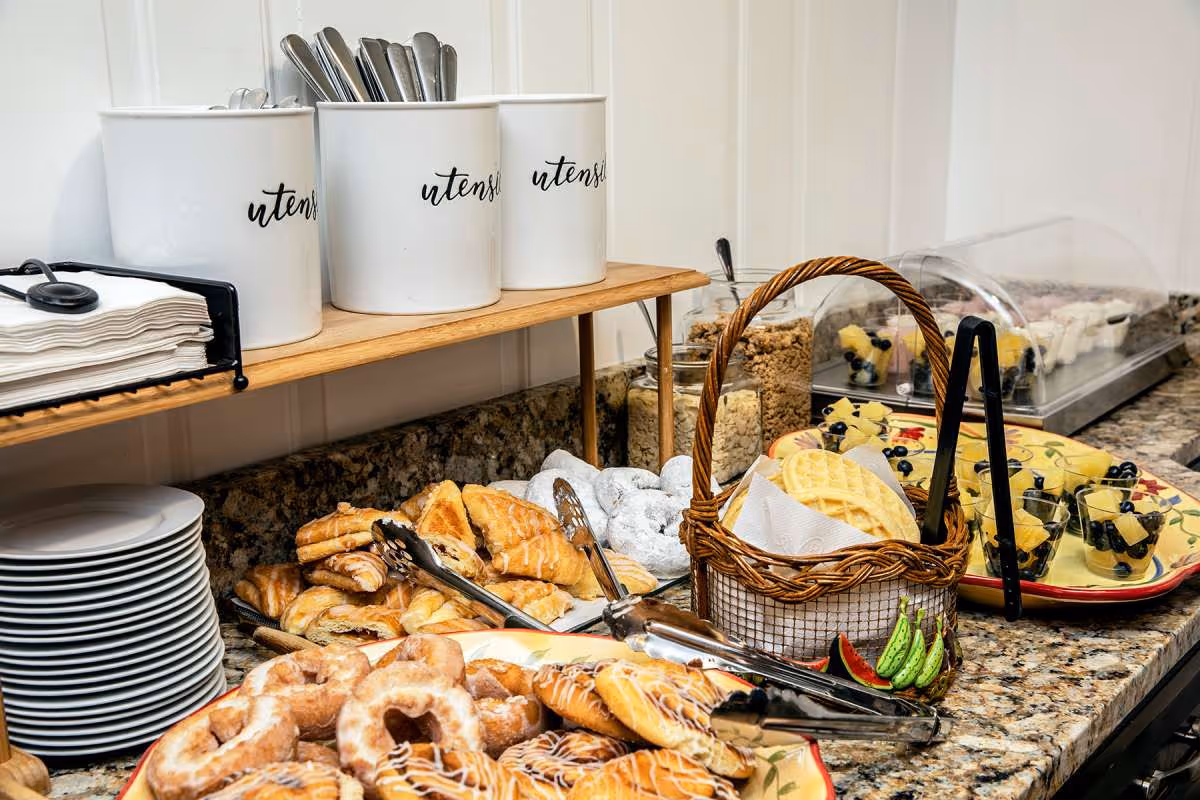 A countertop buffet setup with various breakfast pastries including donuts, waffles, and powdered sugar treats. There are also small cups of fruit and yogurt parfaits, jars of granola and cereal, stacks of plates, napkins, and containers labeled 'utensils' holding silverware.