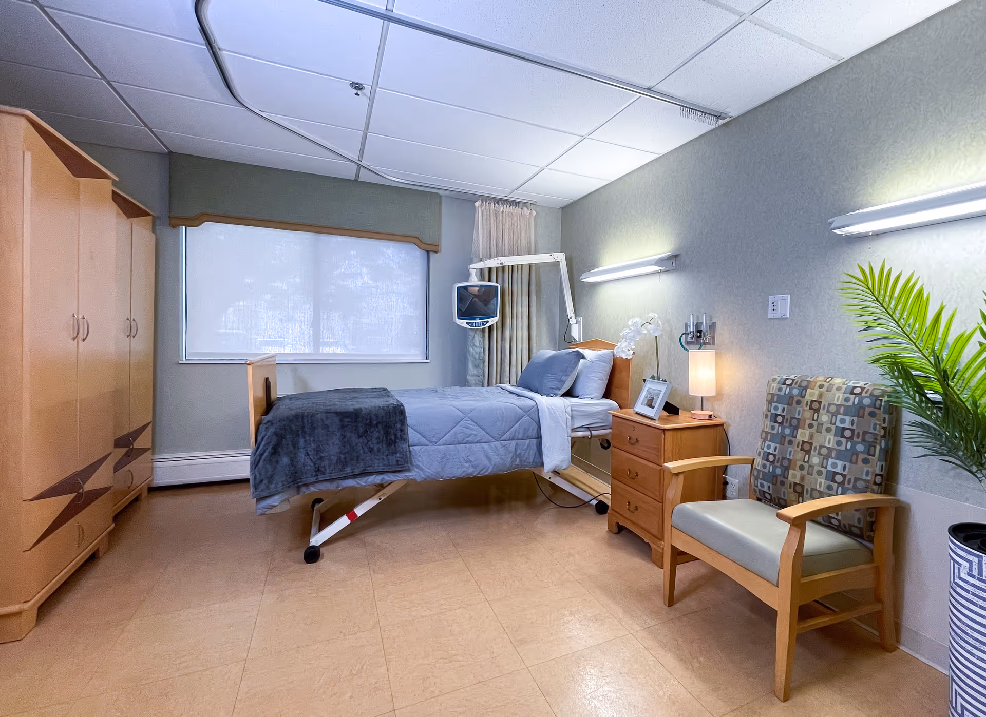 A clean and well-lit bedroom in a health and rehab center featuring a single hospital-style bed with gray bedding and pillows, a wooden nightstand with a lamp and a framed photo, a patterned armchair, a large wooden wardrobe, a potted plant, and a wall-mounted TV. The room has a window with a white blind and soft green walls.