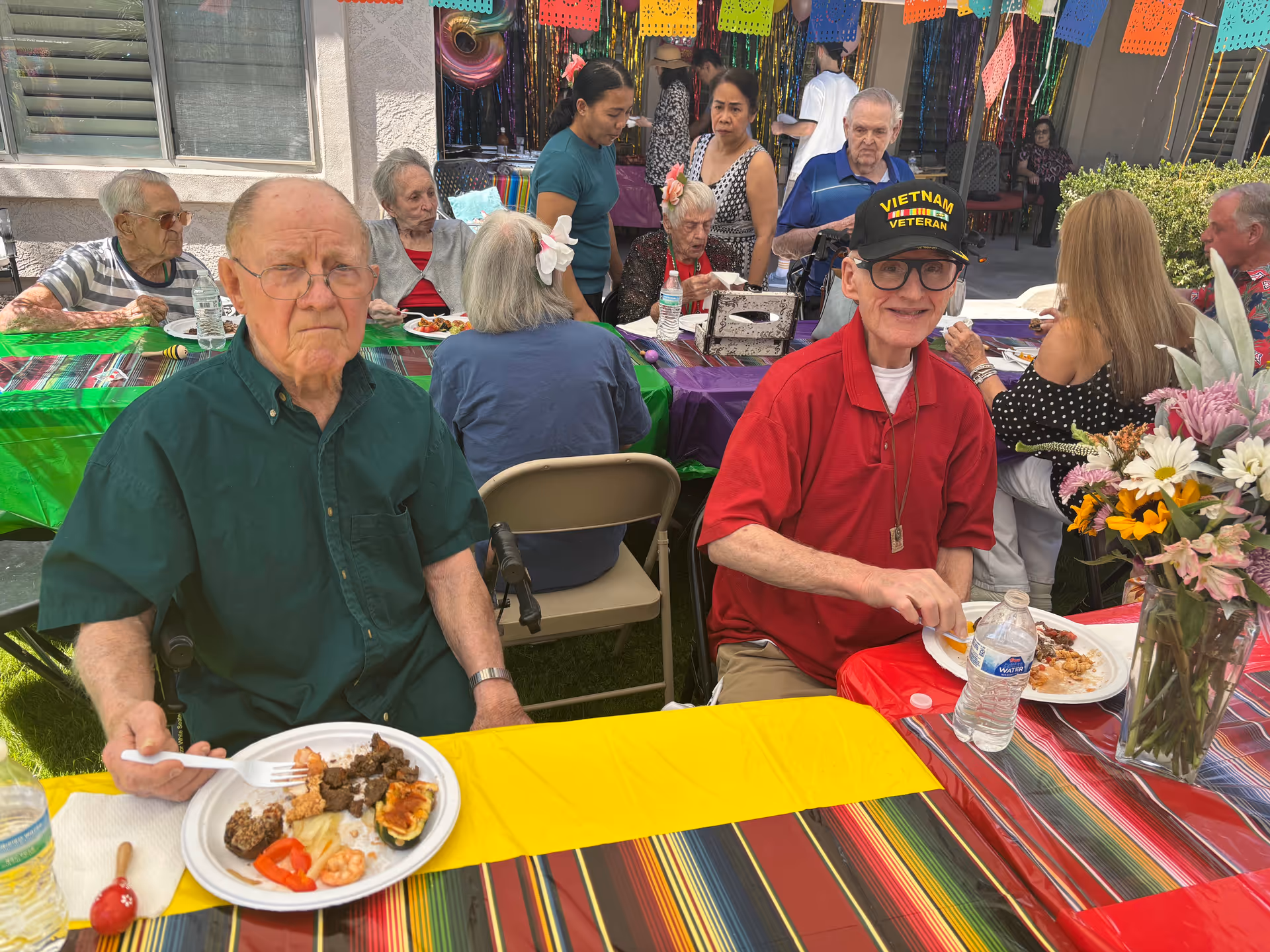 Elderly residents sit at a colorful outdoor dining table during a festive community meal.