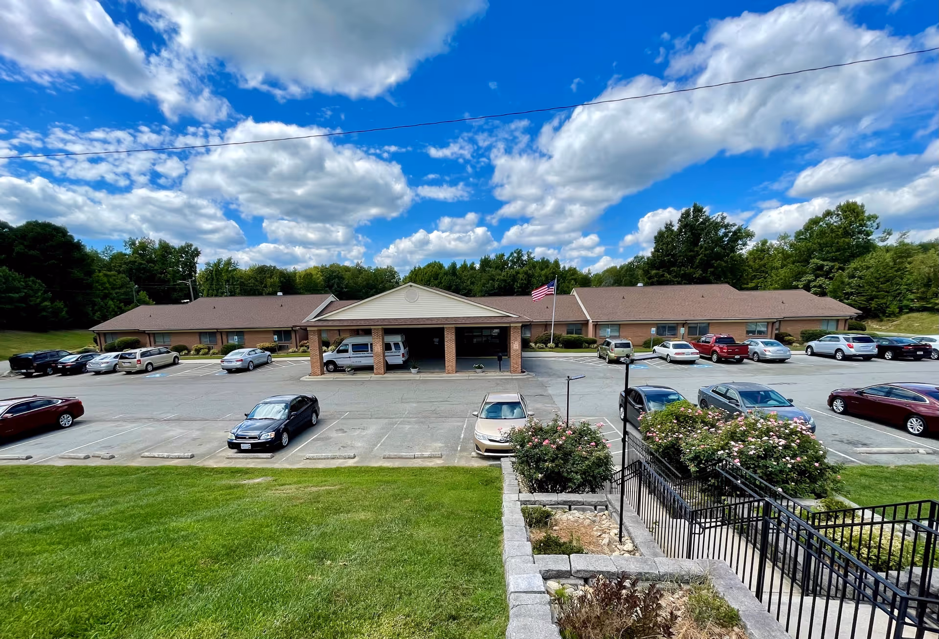 Front exterior view of Roxboro Healthcare & Rehabilitation Center, a single-story brick building with a covered entrance and an American flag on a flagpole. Several cars are parked in the parking lot in front of the building, with green grass and bushes in the foreground and trees in the background under a partly cloudy blue sky.