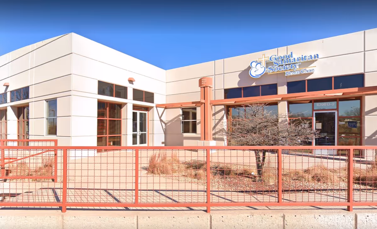 Exterior view of a single-story beige building with a red fence and desert landscaping.