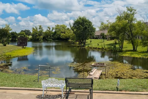 A lakeside outdoor area with a dock, benches, trees, and a grassy lawn.