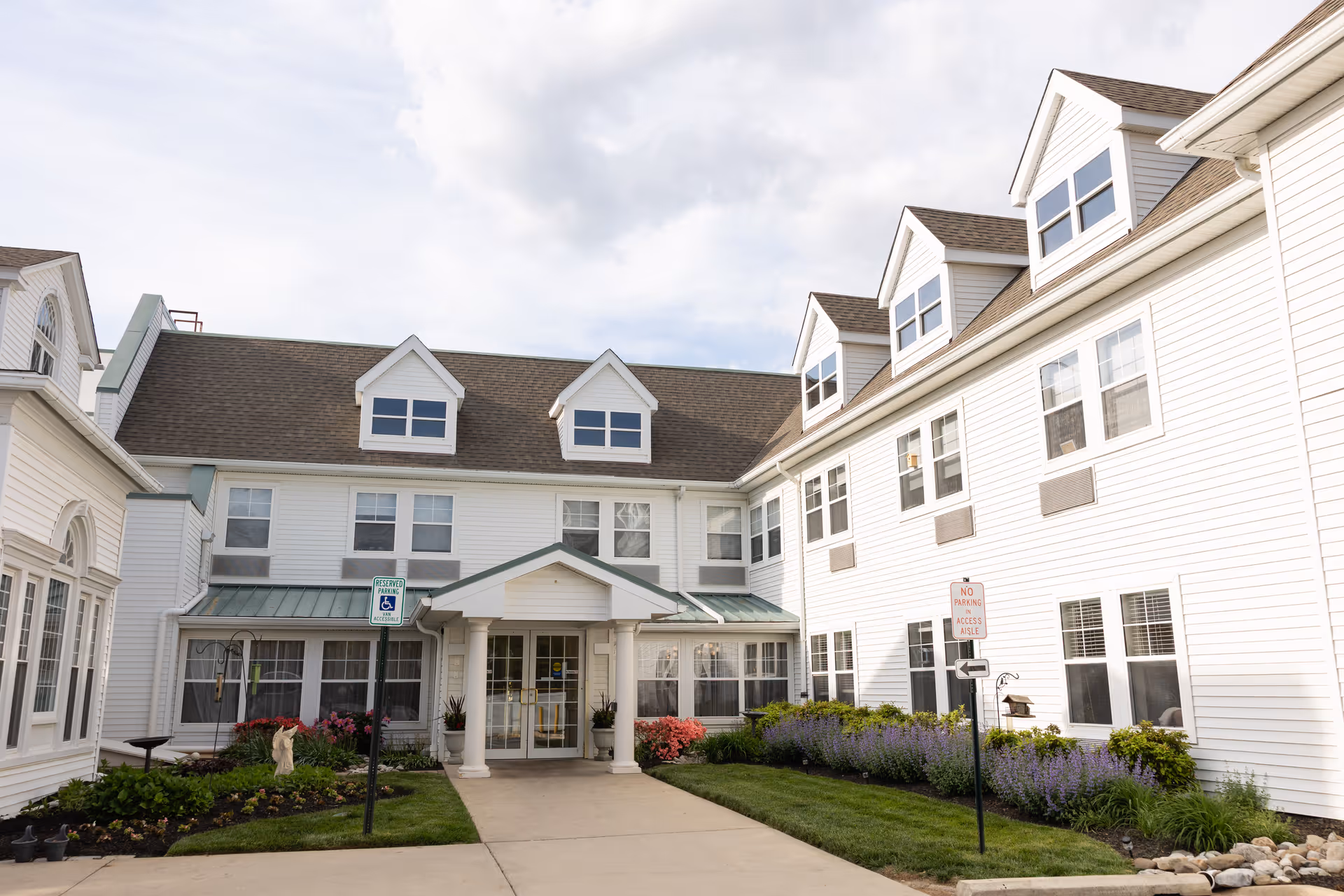 White multi-story building with dormer windows, a covered main entrance and a landscaped walkway.