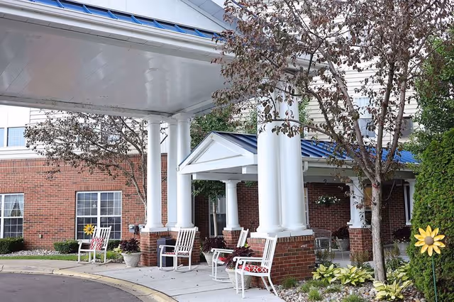 Entrance area of a senior living facility with white columns supporting a covered walkway. There are white rocking chairs with red cushions along the sidewalk, potted plants, and small trees with some greenery and decorative flowers near the building. The building has a brick exterior with white trim and windows.
