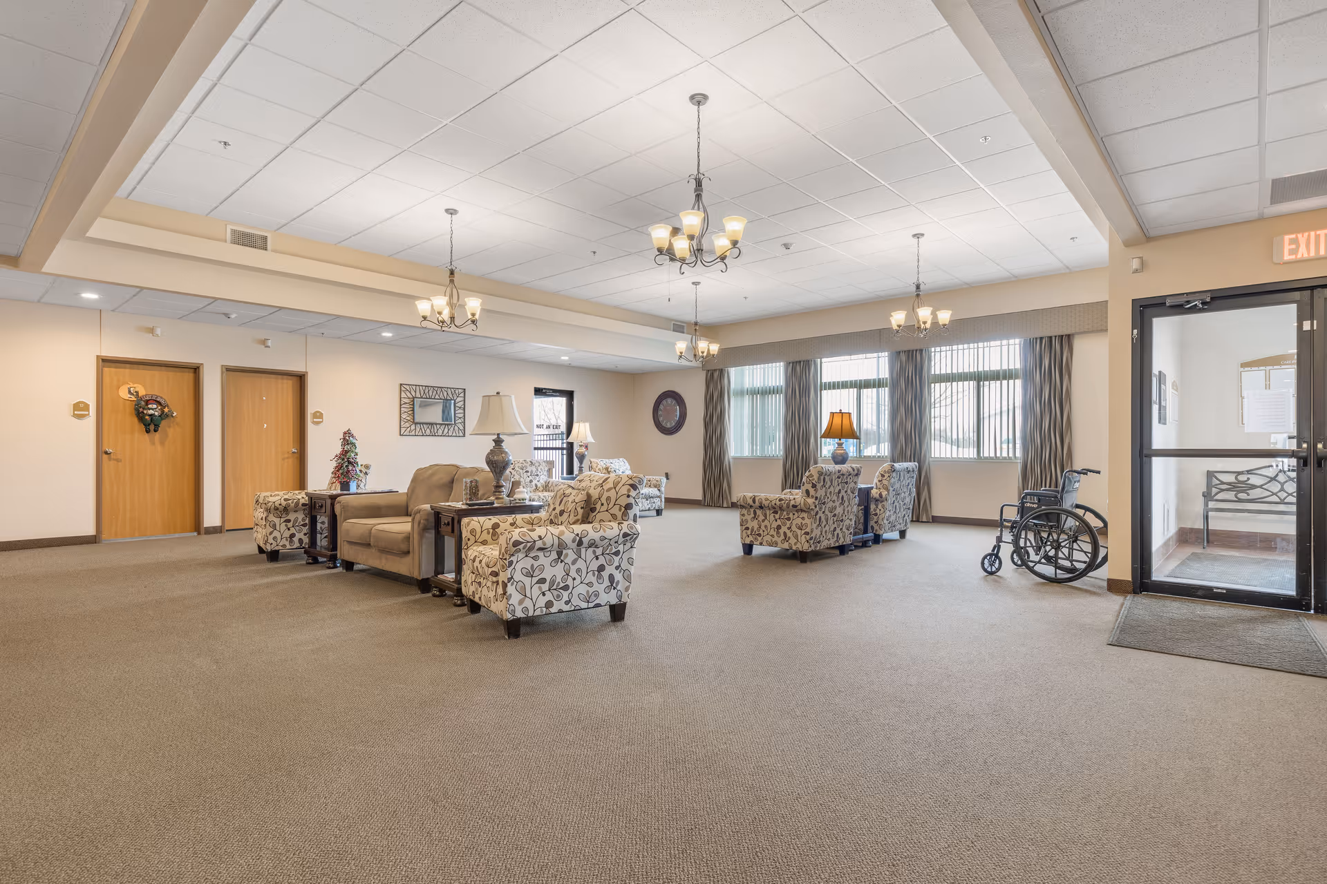 Spacious senior living facility common area with beige carpet, multiple patterned armchairs, a sofa, side tables with lamps, chandeliers hanging from the ceiling, large windows with curtains, a wheelchair near a glass door, and two wooden doors on the left wall.