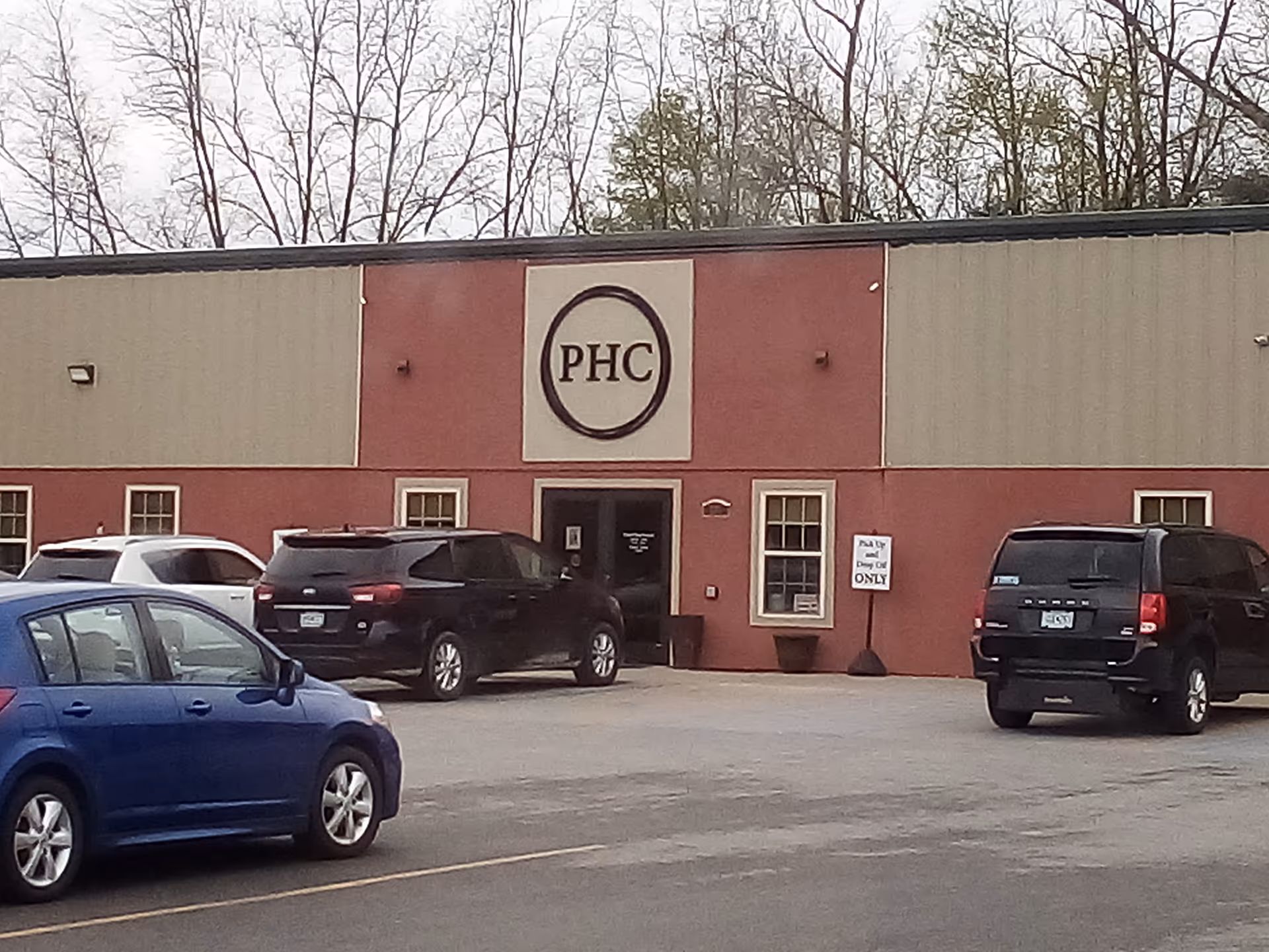 Front view of a red brick facility with a circular PHC logo and parked cars.