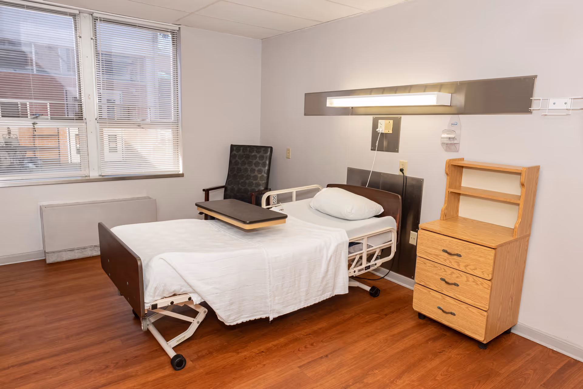 A hospital-style patient room with a bed, dresser, chair, and large window.