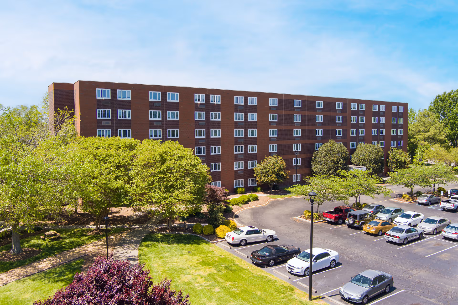Wide exterior view of a multi-story brick residential building with trees, lawn, and a parking lot.