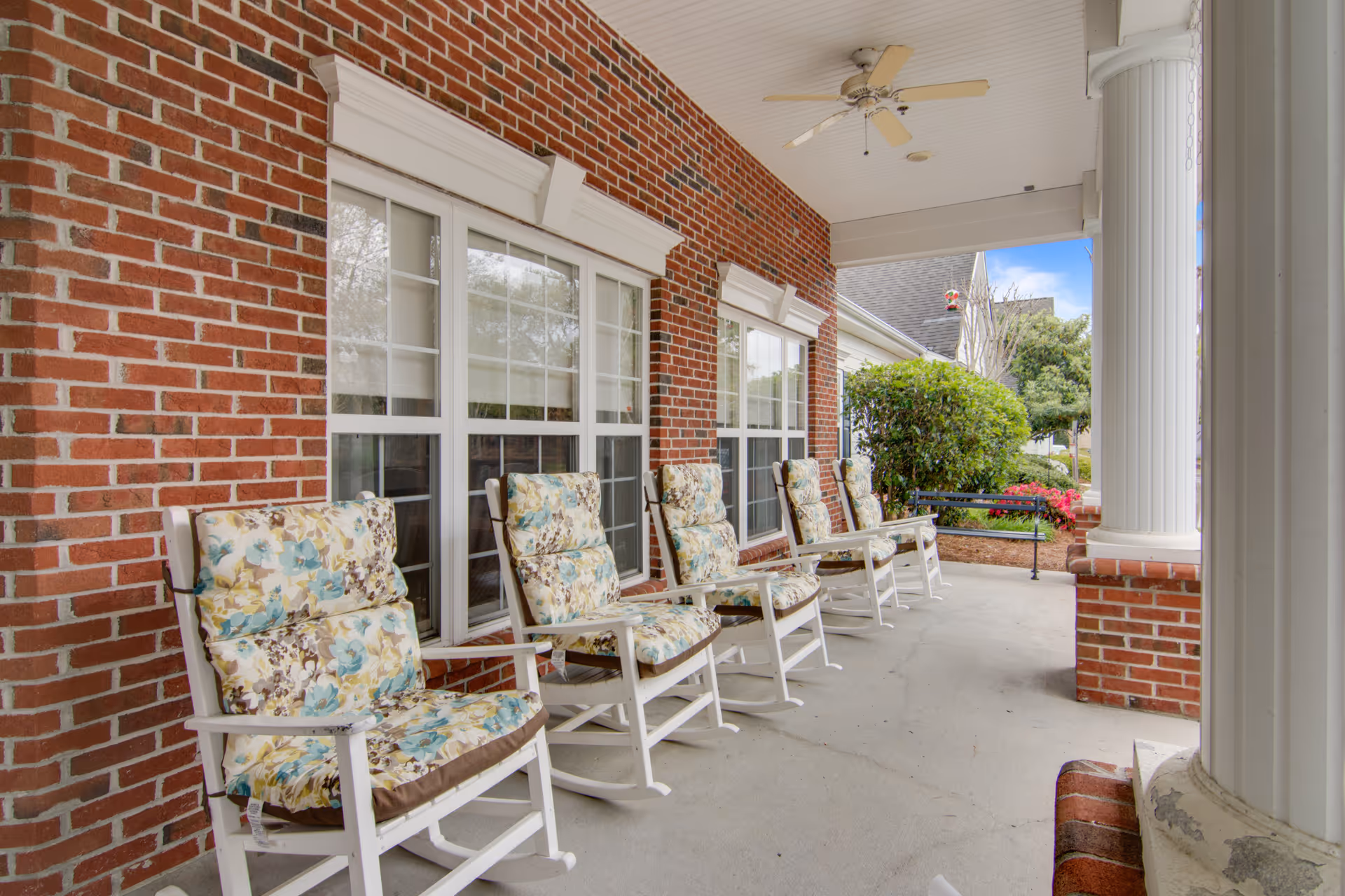 Covered front porch with a row of floral-cushioned white rocking chairs along a brick wall and white columns.