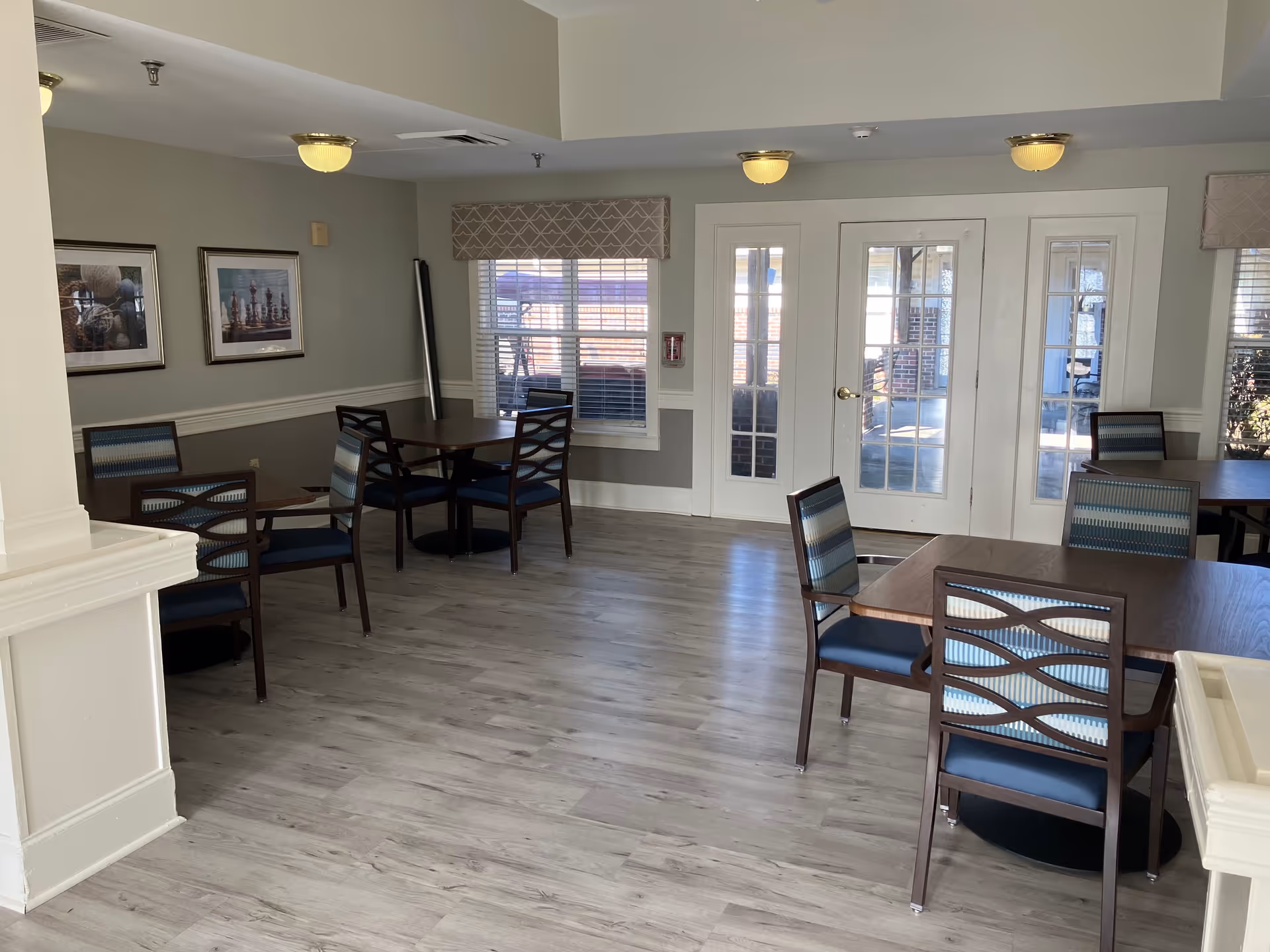 A bright and clean dining area in a senior living facility with several wooden tables and chairs featuring blue patterned cushions. The room has light gray walls, framed artwork, large windows with blinds, and glass double doors leading outside. The floor is light wood laminate.
