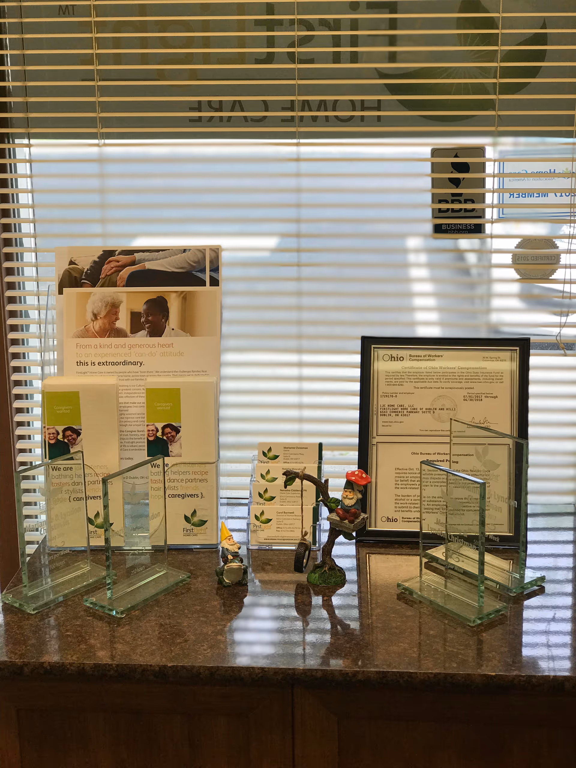 Front desk area with brochures, figurines, and a framed certificate behind blinds in a lobby.