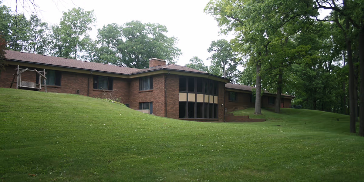 Brick senior living facility on a grassy hill, surrounded by tall trees and large windows.