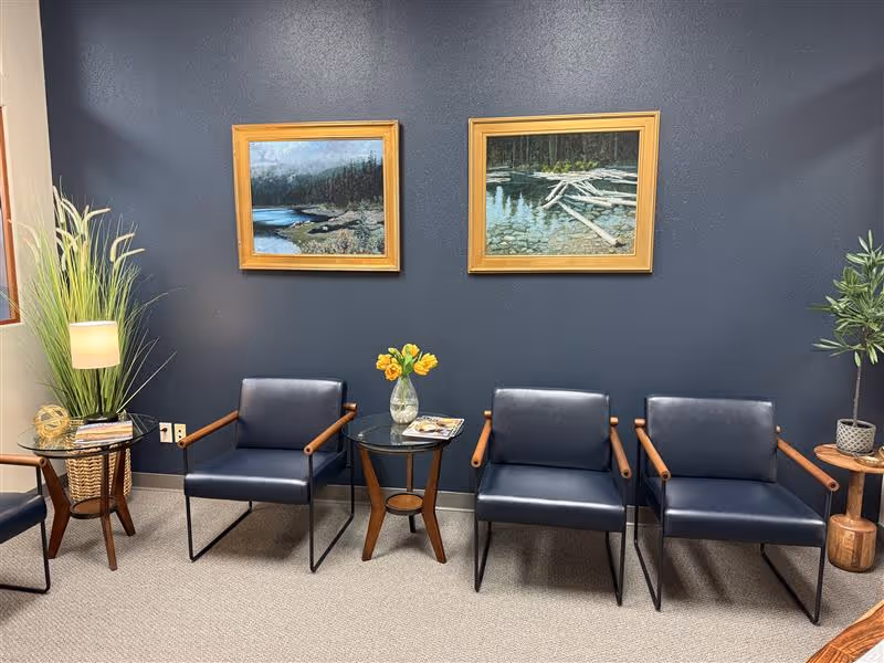Indoor waiting area with blue chairs, framed pictures, plants, and a glass table.