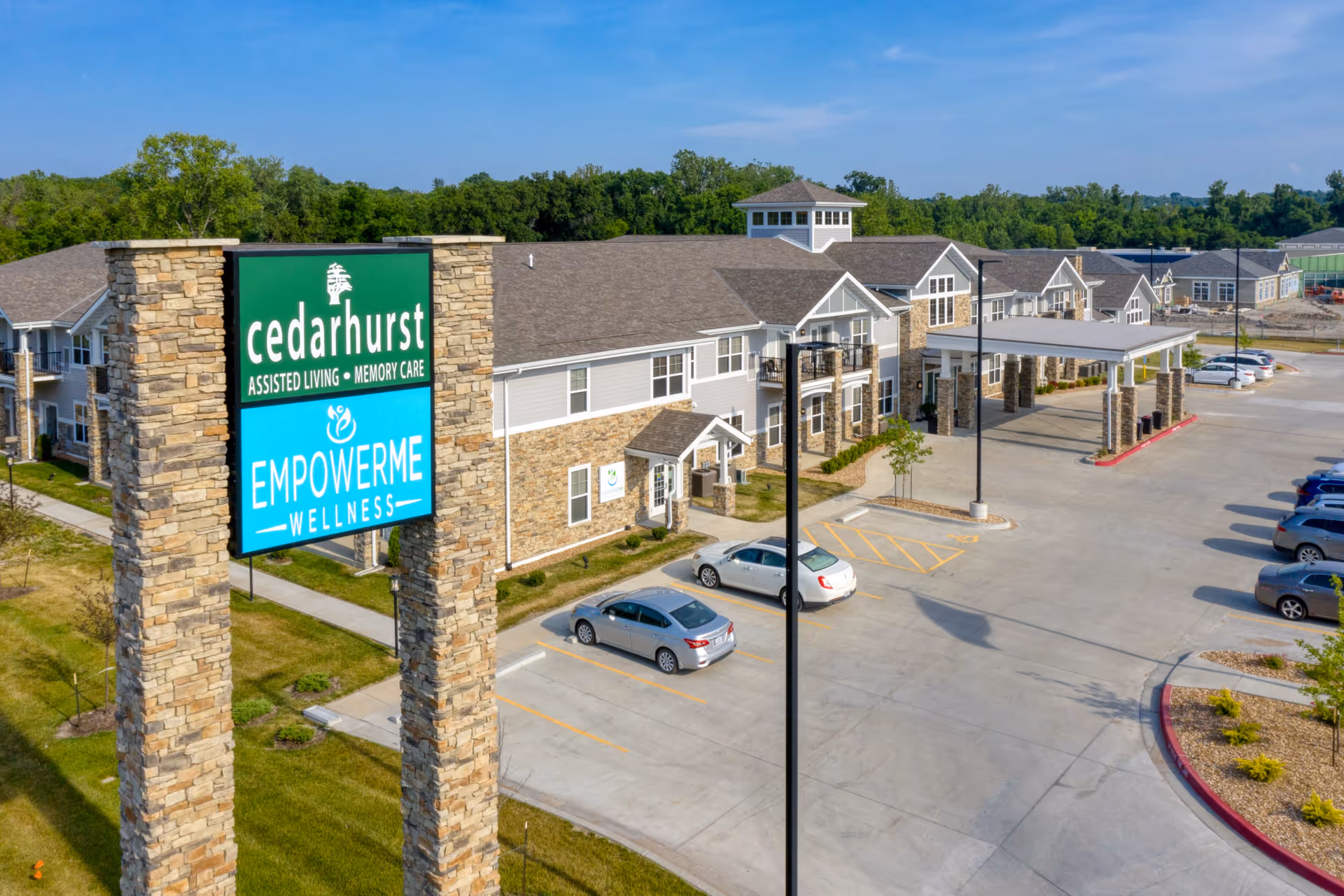 Exterior view of Cedarhurst Senior Living of Blue Springs building with a large stone sign in the foreground displaying the facility name and services. The building is two stories with a covered entrance and a parking lot with several cars. Trees and greenery surround the area under a clear blue sky.