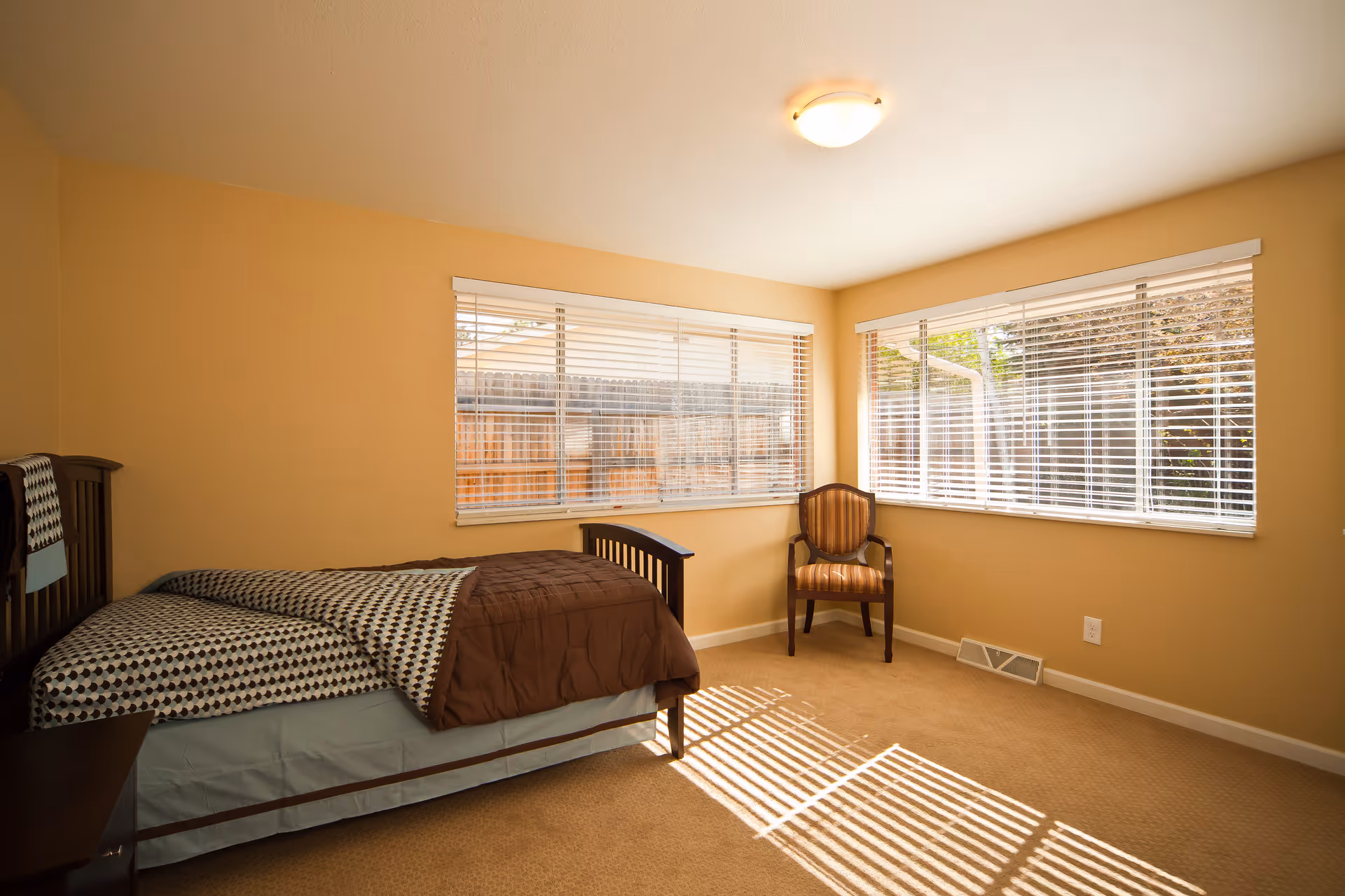 Sunlit bedroom with a bed, chair, and large windows with blinds.