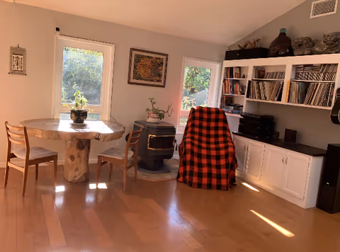 Sunlit living room with a round wooden table and chairs, a plaid-covered armchair, a wood stove, and built-in shelving with books and records.