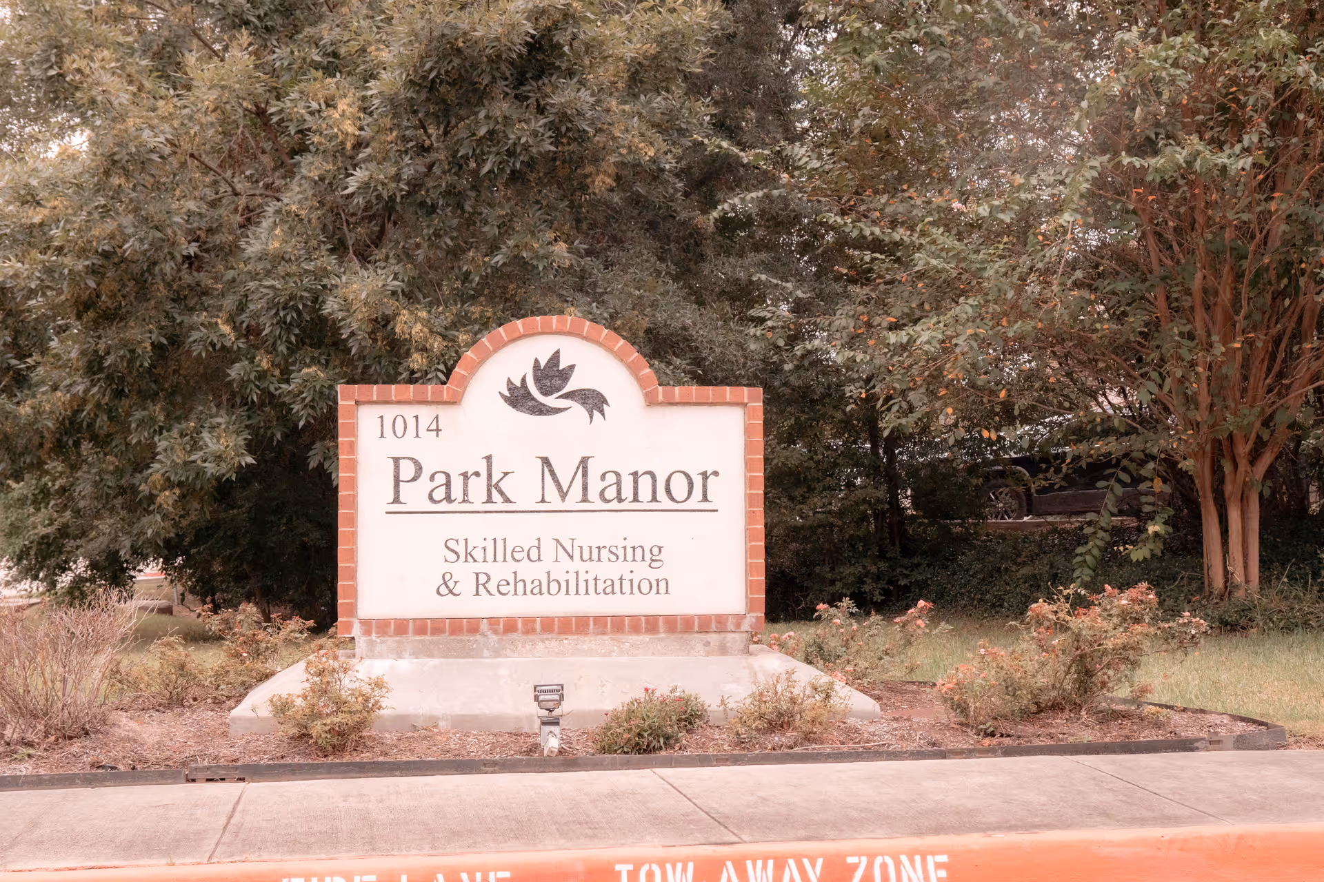 A brick and concrete sign for Park Manor Skilled Nursing & Rehabilitation, located at 1014, surrounded by bushes and trees with a sidewalk and a red curb in front.