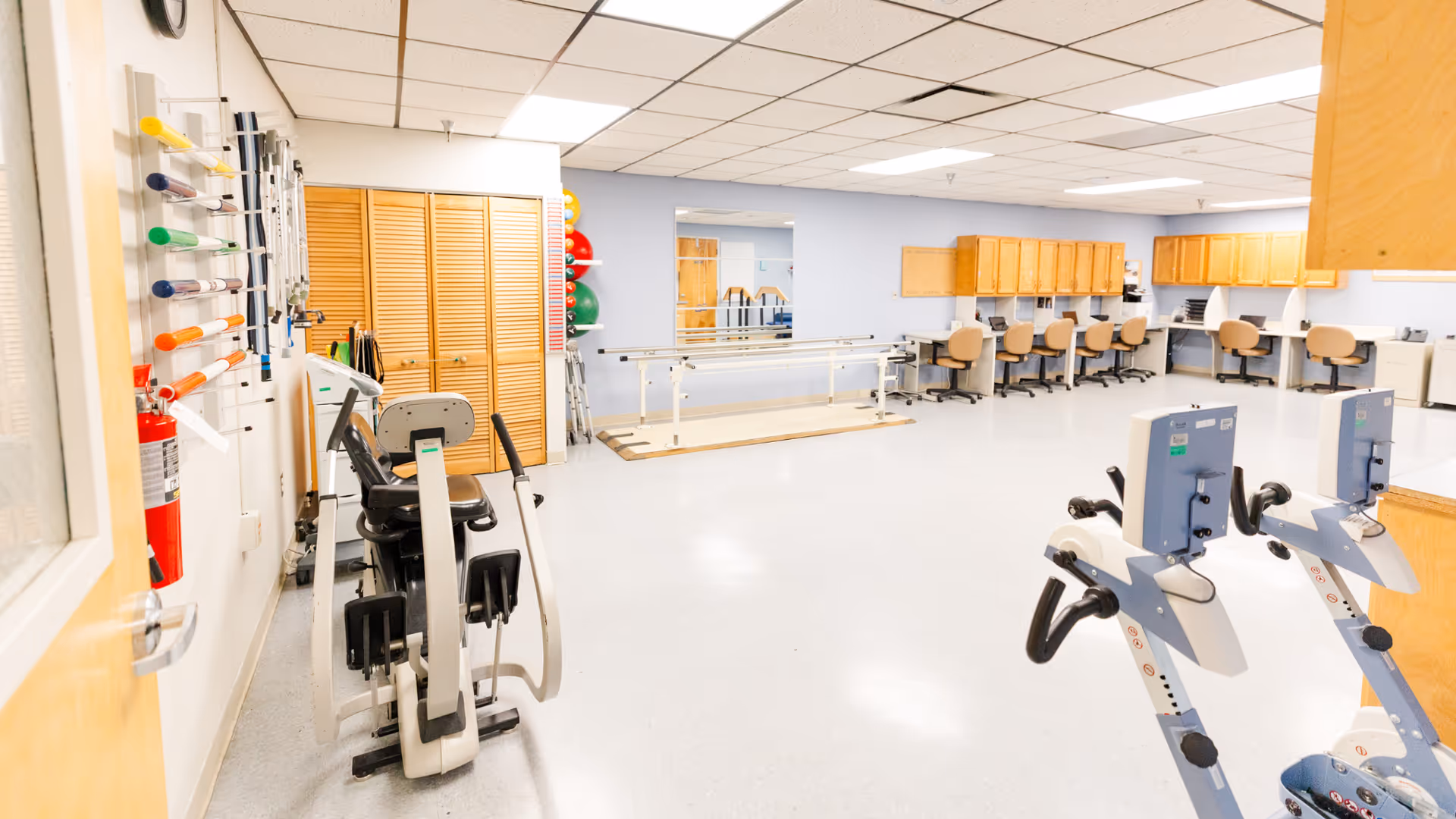 Bright rehabilitation gym with exercise bikes, therapy equipment, and a row of workstations along back.