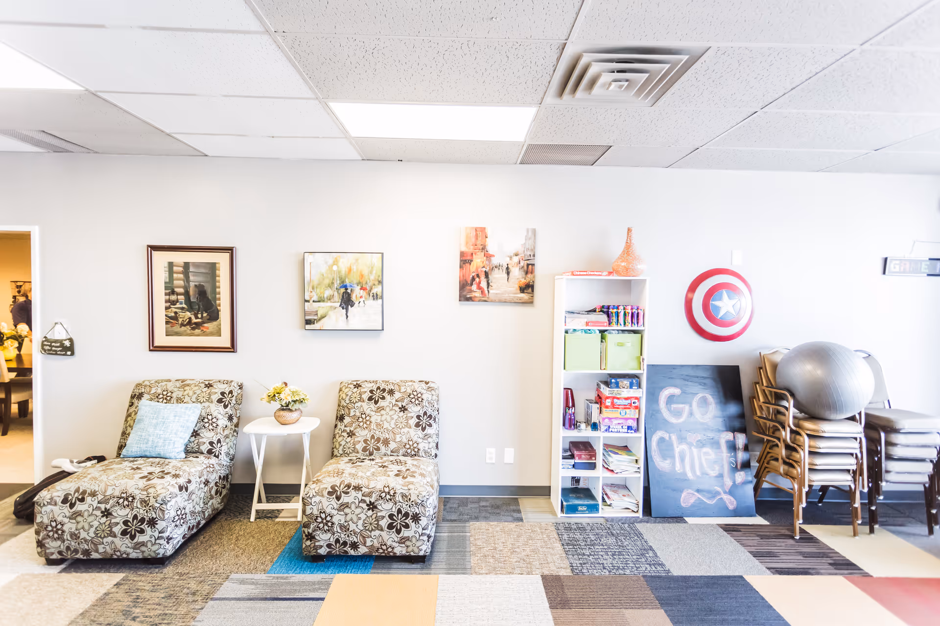 Cozy lounge area with patterned chairs, small table, wall art, shelves, and exercise gear.