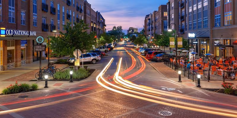 Nighttime downtown street with lit storefronts, light trails, parked cars, and outdoor seating along the sidewalk.
