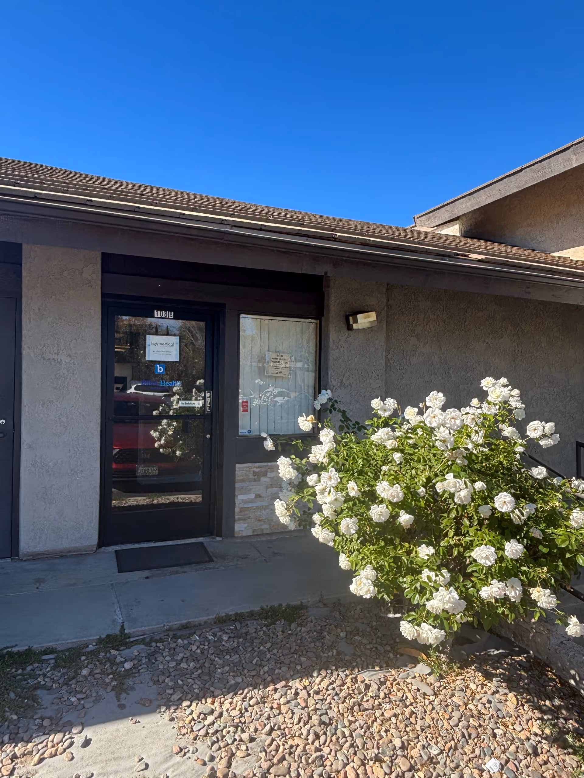 Exterior of a facility entrance with glass door, windows, and a white flowering shrub.