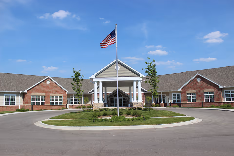 Front exterior view of a single-story senior living facility building with a circular driveway, a flagpole with an American flag in the center, and a covered entrance supported by white columns. The building has a combination of brick and siding exterior with multiple windows and a clear blue sky in the background.