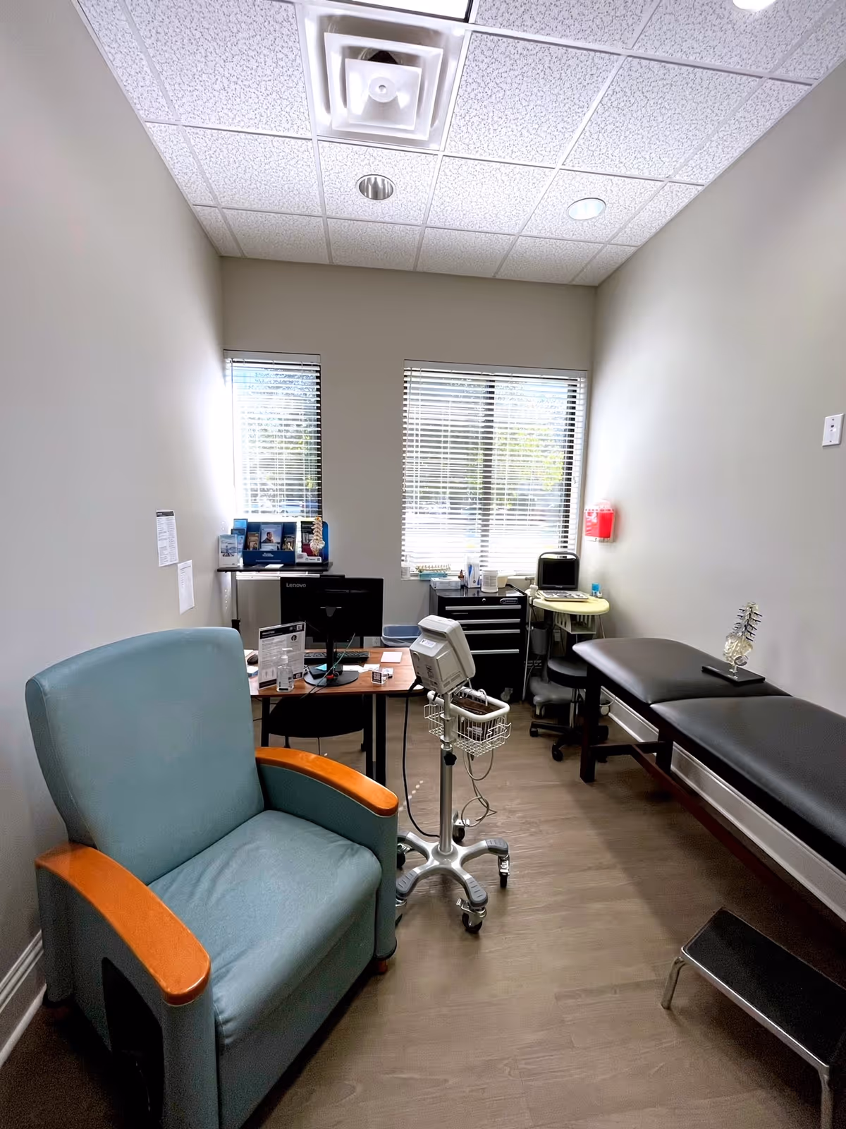 Small medical examination room with teal armchair, desk, computer, and examination bed.