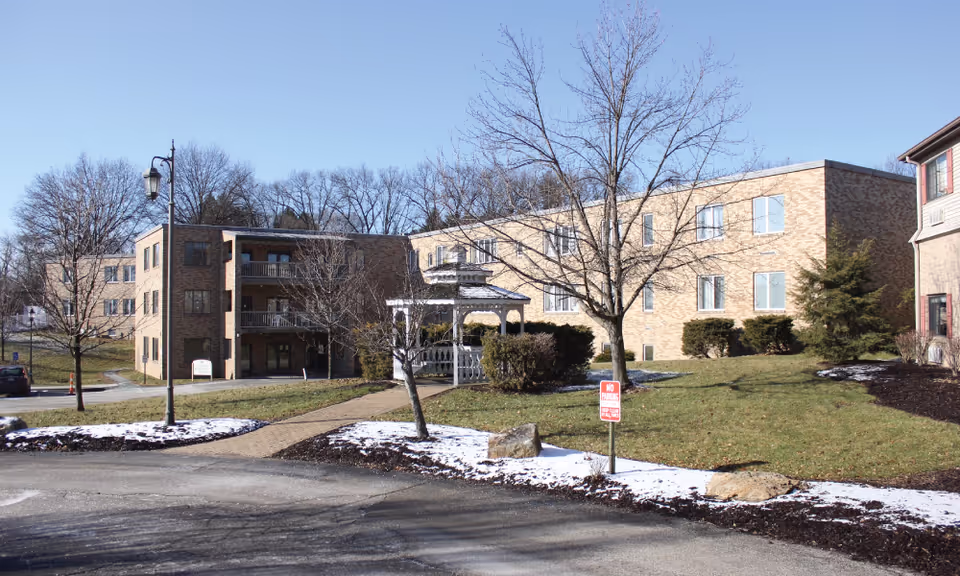 Front exterior of a brick senior living building with a white gazebo, leafless trees, and patches of snow on the lawn.