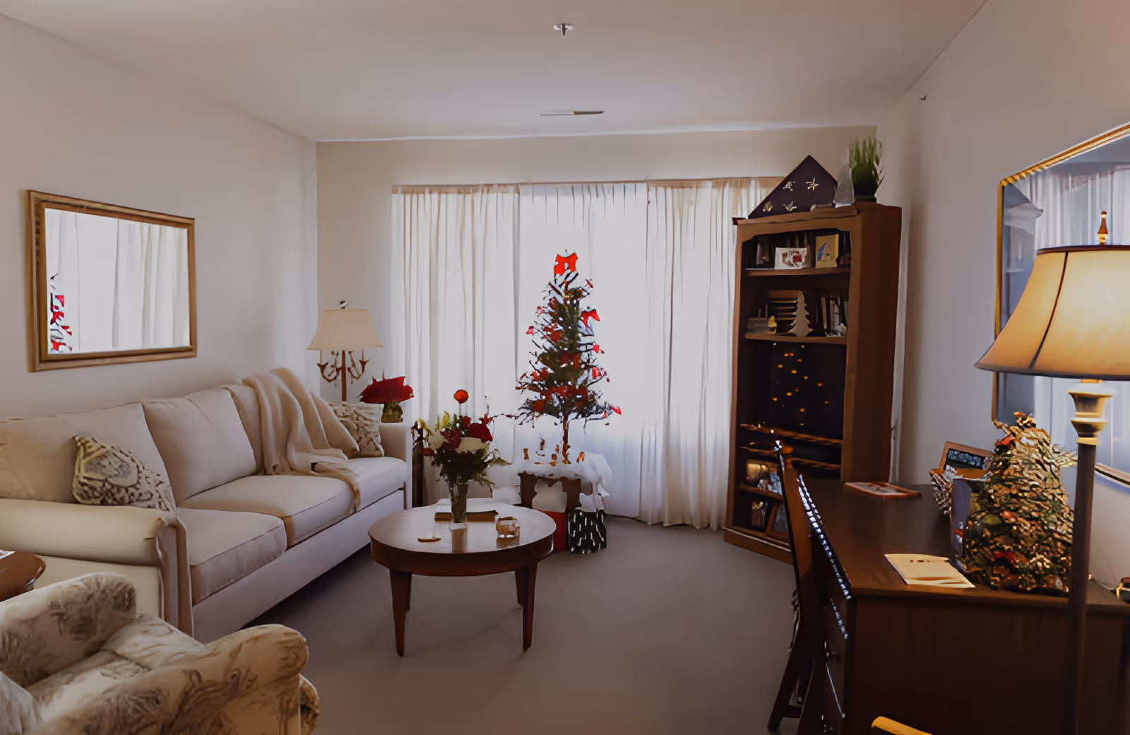 A cozy living room with a beige sofa, an armchair, a wooden coffee table with a vase of flowers, a small decorated Christmas tree near a window with white curtains, a wooden bookshelf with various items, and a wooden desk with a lamp and decorative objects.