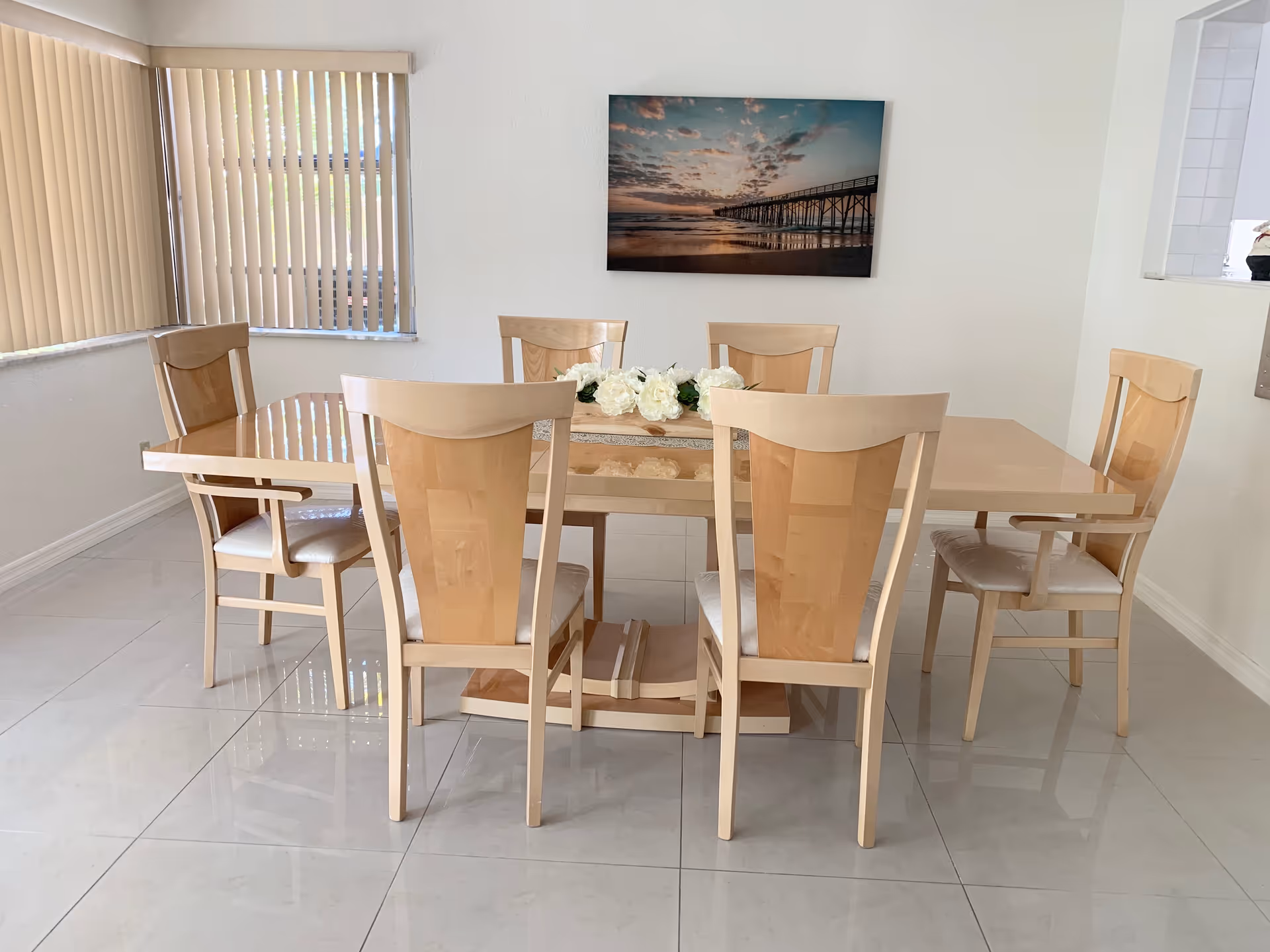 A bright dining room with a wooden table, six chairs, blinds, and wall art.