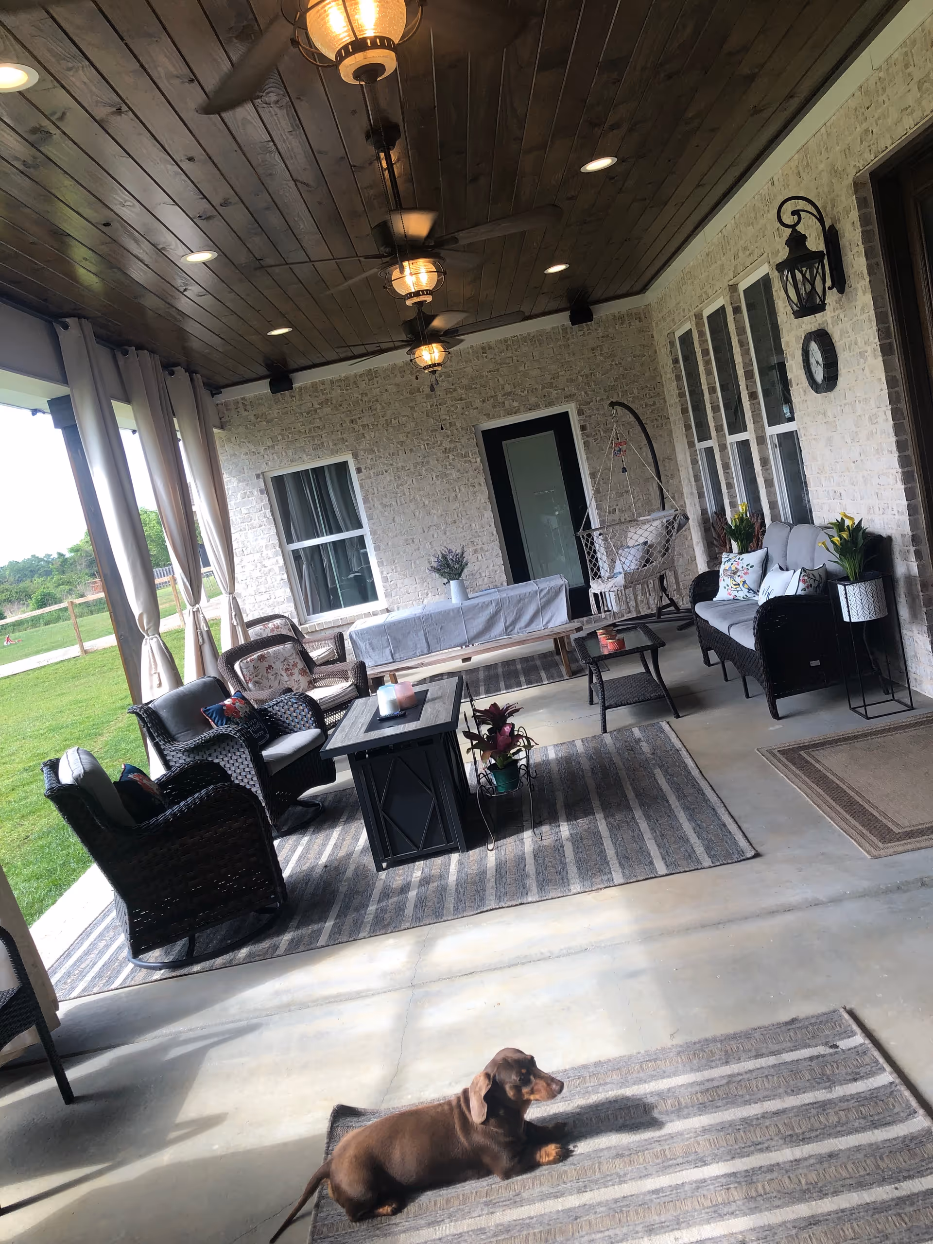 Covered outdoor patio with wicker seating, rugs, plants, ceiling fans, and a small dog.