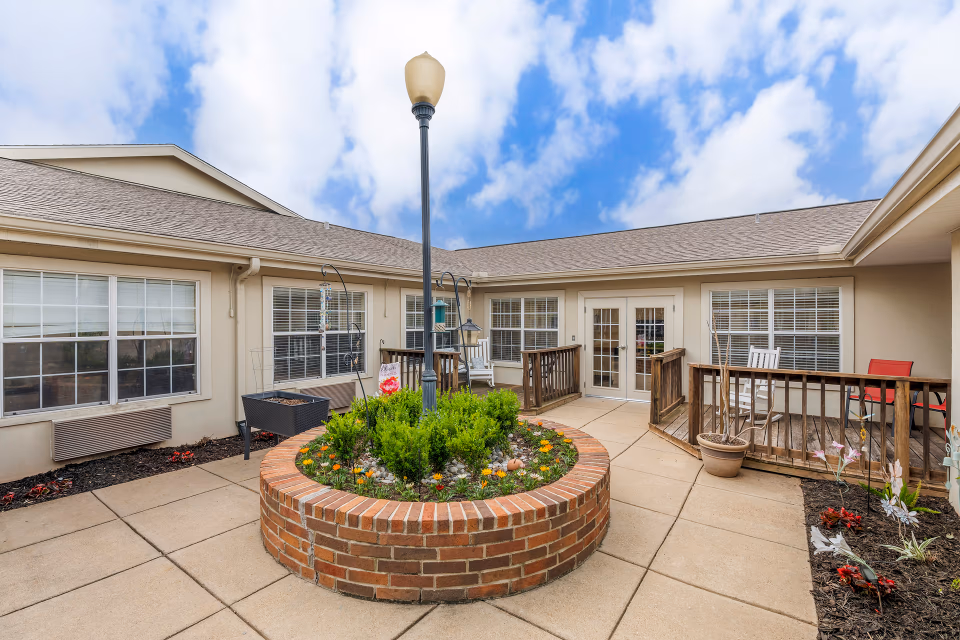 Outdoor courtyard area with a circular brick planter filled with greenery and flowers, a tall lamppost in the center, and a building with multiple windows and a double door surrounding the courtyard. There are wooden railings and some outdoor chairs on the patio.